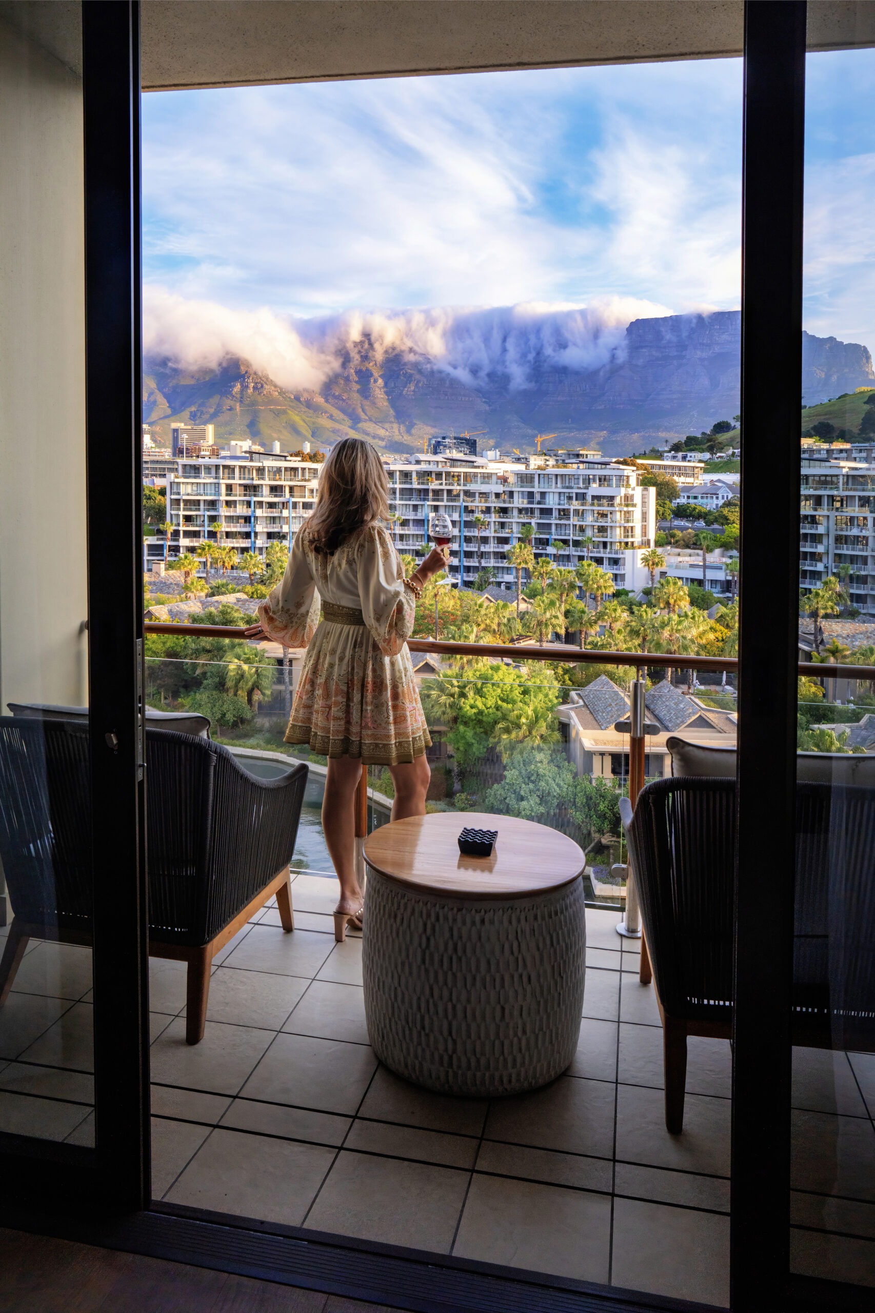 A woman stands on the balcony of the Presidential Suite at the One&Only Cape Town, enjoying a sunset cocktail with Table Mountain draped in clouds in the background — luxury family travel photography by David Scarola.David Scarola Photography, One and Only Cape Town Presidential Suite, luxury family travel photographer, family photography Cape Town, global family photo shoots, destination family portraits, high-end event photography South Africa, Table Mountain sunset view, lifestyle travel photography, luxury resort photography, global event coverage photographer, international family photographer, editorial travel photographer, Cape Town balcony sunset photo.