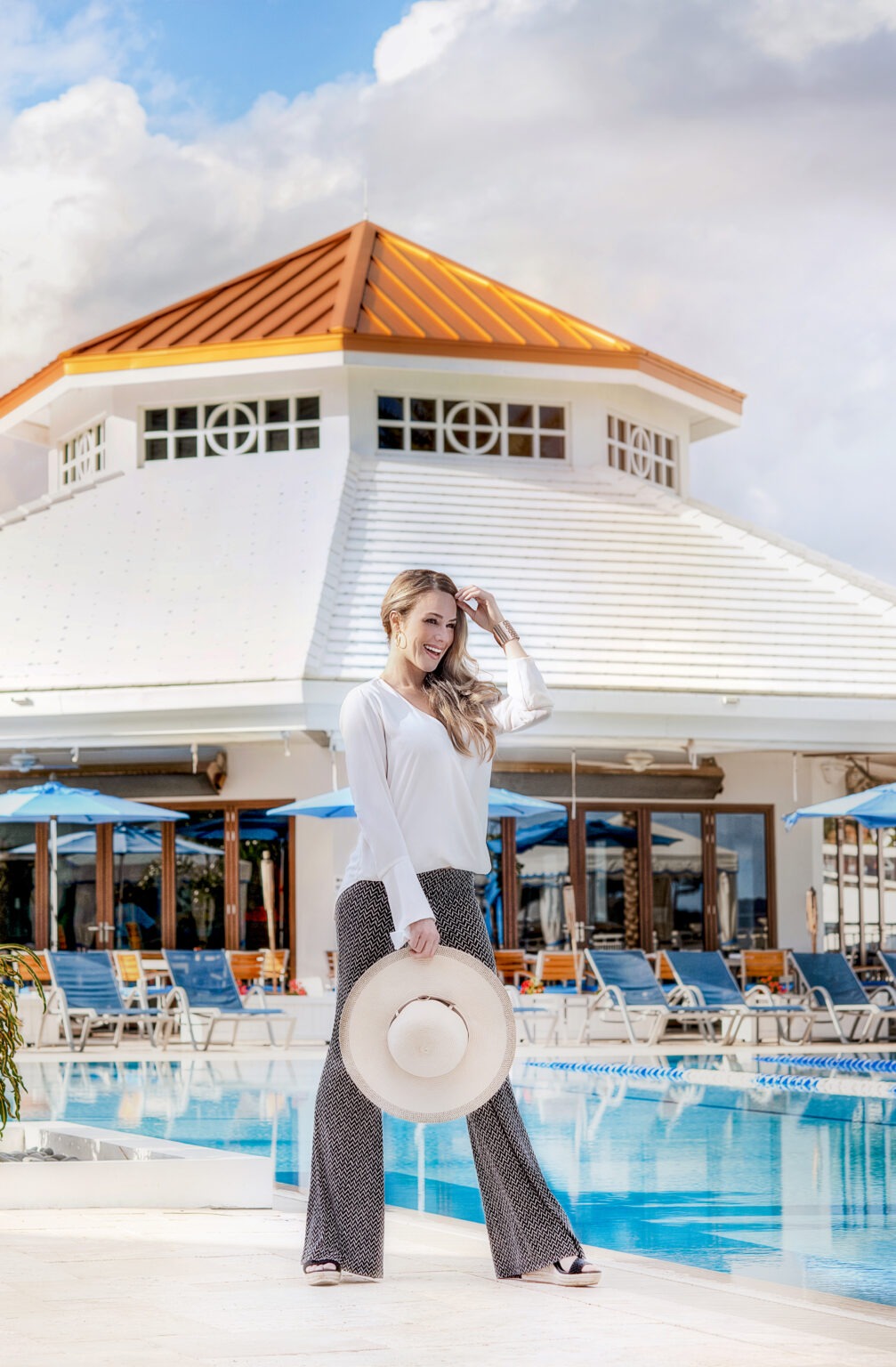 Luxury fashion photograph featuring a model wearing Ceci Palm Beach resort wear during a high-end catalogue shoot at the Admirals Cove Country Club in Jupiter Island, Florida, captured by David Scarola, one of the leading fashion and lifestyle photographers in Palm Beach. The model, dressed in a white blouse, patterned palazzo pants, and holding a wide-brimmed hat, stands confidently by the pool under bright blue skies — representing timeless Palm Beach sophistication and coastal chic style.