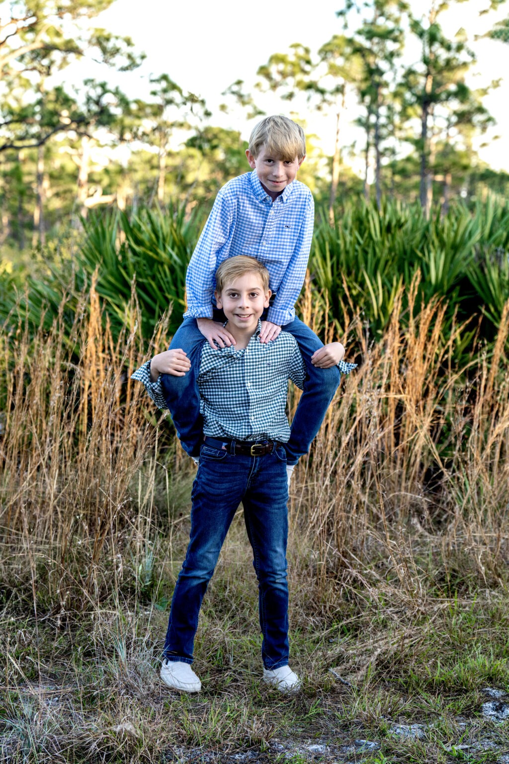 Two brothers, Grady and Gunnar, share a joyful moment during a family photo shoot in the palm tree forest of Jupiter, Florida. The boys, dressed in coordinated plaid shirts and jeans, capture the essence of brotherly love and adventure surrounded by natural Florida greenery. Photographed by David Scarola Photography, renowned for fine art family portraiture in Palm Beach County.family photographer Jupiter FL, Palm Beach family portraits, brothers photoshoot Florida, sibling photography Jupiter Island, lifestyle photographer Palm Beach County, outdoor family photo ideas, David Scarola Photography, best Jupiter Florida photographer, natural light family photographer, Palm Beach lifestyle portraits.