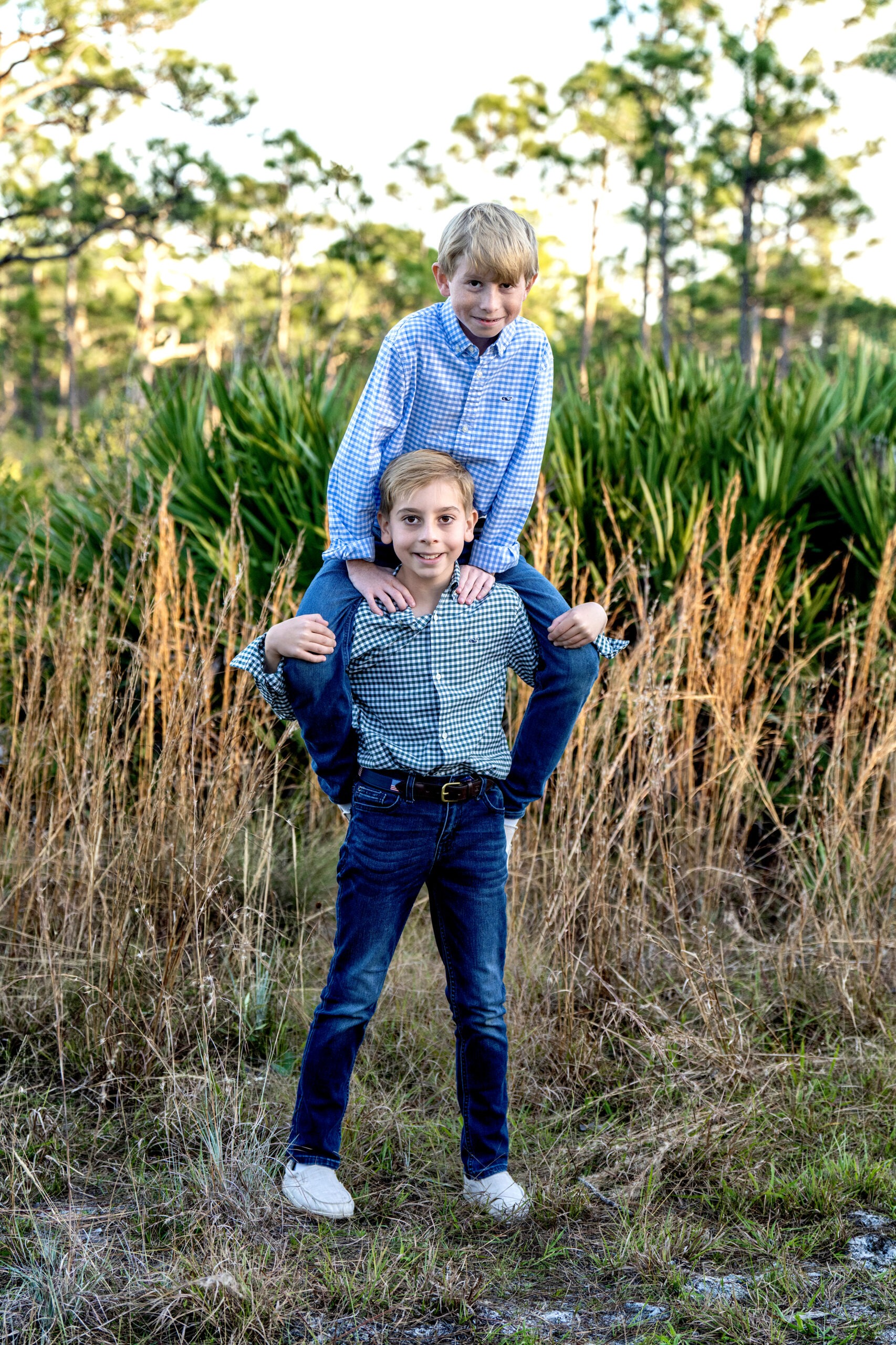 Two brothers, Grady and Gunnar, share a joyful moment during a family photo shoot in the palm tree forest of Jupiter, Florida. The boys, dressed in coordinated plaid shirts and jeans, capture the essence of brotherly love and adventure surrounded by natural Florida greenery. Photographed by David Scarola Photography, renowned for fine art family portraiture in Palm Beach County.family photographer Jupiter FL, Palm Beach family portraits, brothers photoshoot Florida, sibling photography Jupiter Island, lifestyle photographer Palm Beach County, outdoor family photo ideas, David Scarola Photography, best Jupiter Florida photographer, natural light family photographer, Palm Beach lifestyle portraits.