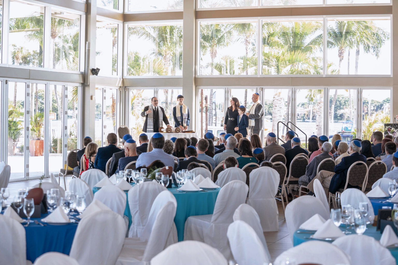 Bar Mitzvah ceremony overlooking the Intracoastal Waterway at The Lake Pavilion in Palm Beach, Florida. Guests gather in a bright, sunlit ballroom as the rabbi leads the family through sacred readings before the Torah, surrounded by panoramic windows and palm trees.Bar Mitzvah photographer Palm Beach, luxury event photographer Florida, The Lake Pavilion Palm Beach events, Jewish family celebrations, private event photography, high-end religious ceremony photography, destination event photographer, David Scarola Photography, top Palm Beach event photographer, global private family event photography.