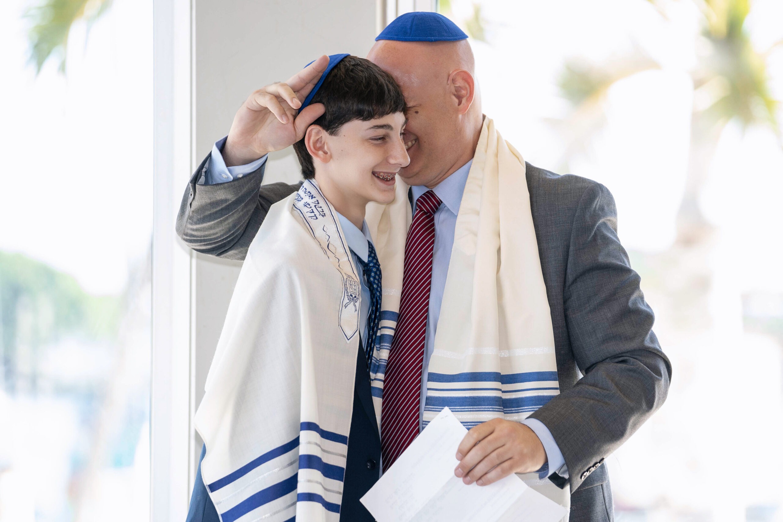 A touching Bar Mitzvah moment between father and son at The Lake Pavilion in Palm Beach, Florida. The two share a heartfelt embrace, wrapped in traditional tallit prayer shawls, symbolizing faith, family, and Jewish heritage.Bar Mitzvah photographer Palm Beach, luxury family event photography, Lake Pavilion Palm Beach, Jewish event photographer Florida, worldwide private event photography, Jupiter Island family photographer, high-end religious photography, David Scarola Photography, top-rated event photographer, father son Bar Mitzvah moment.