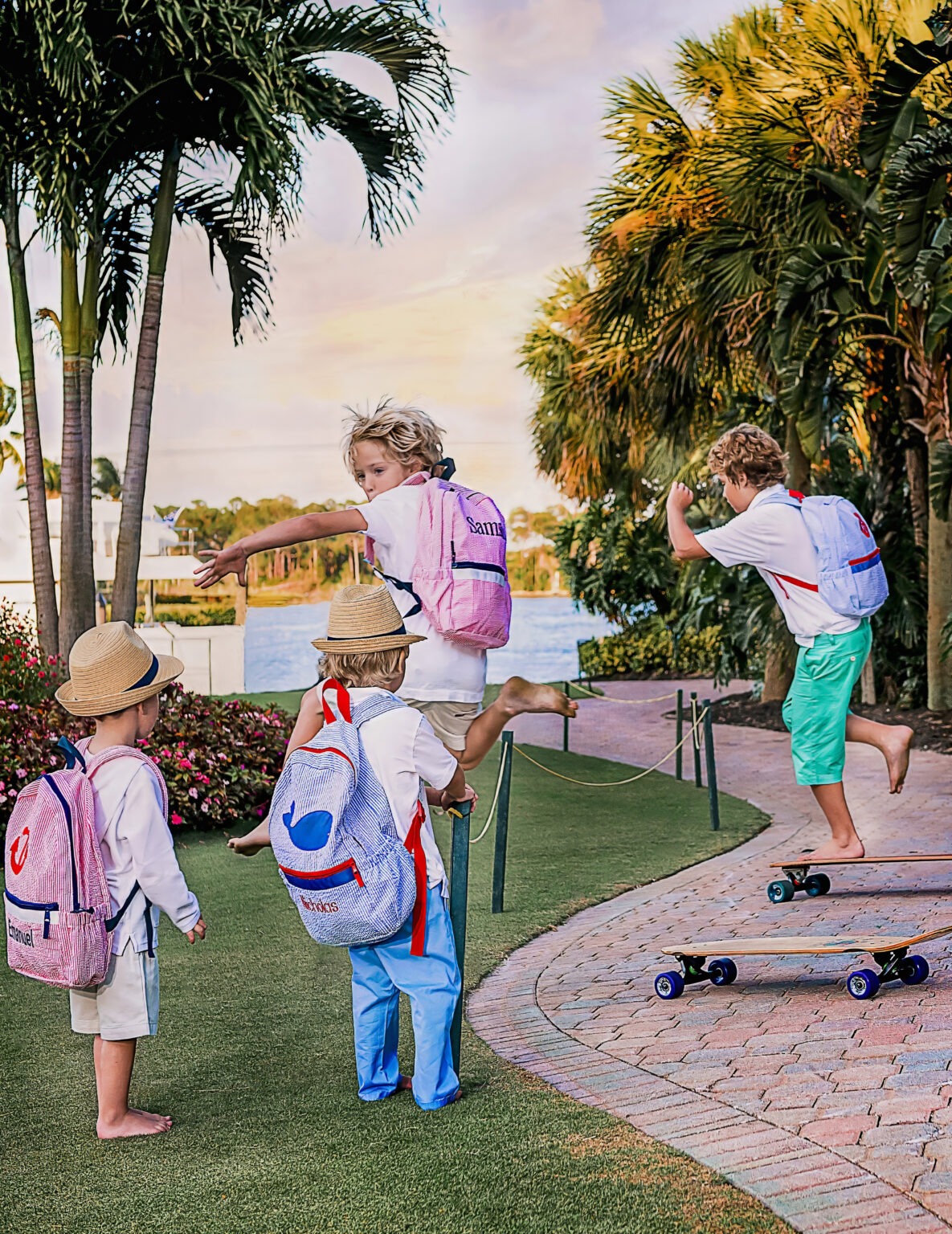 Four young boys wearing colorful Palm Beach Crew backpacks from Nikiani Inc. play and skateboard at sunset along the waterfront at Admirals Cove Country Club in Jupiter, Florida. Captured by David Scarola Photography.
