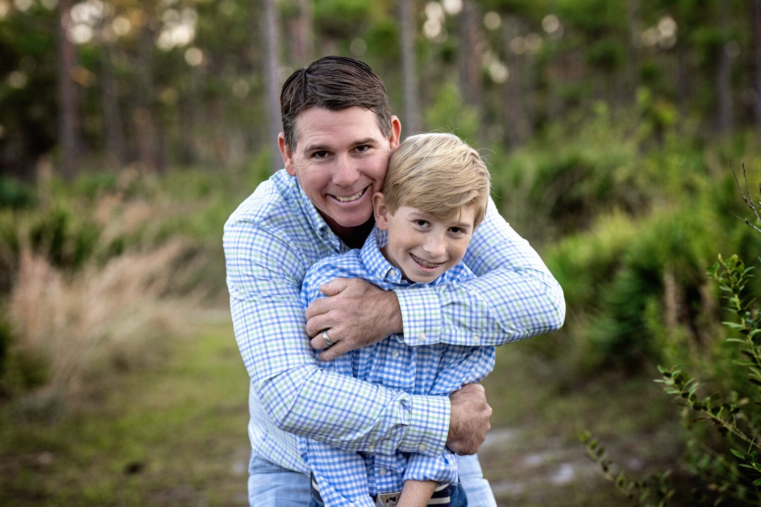 A father and son share a joyful embrace during a family photography session in the pine tree forest of Jupiter, Florida. Dressed in matching plaid shirts, they radiate warmth, connection, and love. Photographed by David Scarola Photography, master of fine art and lifestyle family portraits in Palm Beach and Jupiter Island.family photographer Jupiter FL, Palm Beach family photography, father and son photo shoot, outdoor portraits Jupiter Island, pine forest photography Florida, lifestyle photographer Palm Beach County, David Scarola Photography, best family photographer South Florida, fine art family portraits, candid family photography Jupiter.