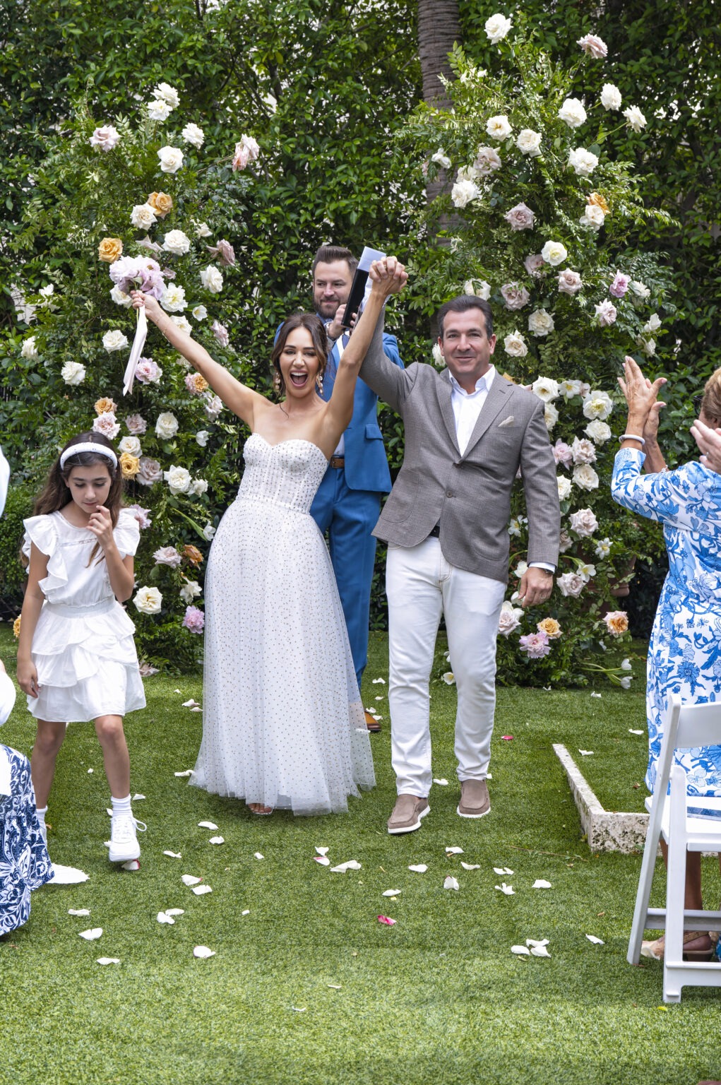 Newly married couple celebrates under a lush floral arch at The Colony Hotel in Palm Beach — the bride radiant in a modern crystal-studded gown, bouquet raised in pure triumph, the groom smiling with quiet grace beside her. Guests cheer, petals fall, and joy fills the garden. Captured by David Scarola, a Palm Beach fine-art wedding photographer known for transforming fleeting emotion into cinematic imagery — blending light, texture, and story with painterly precision and soulful timing.
