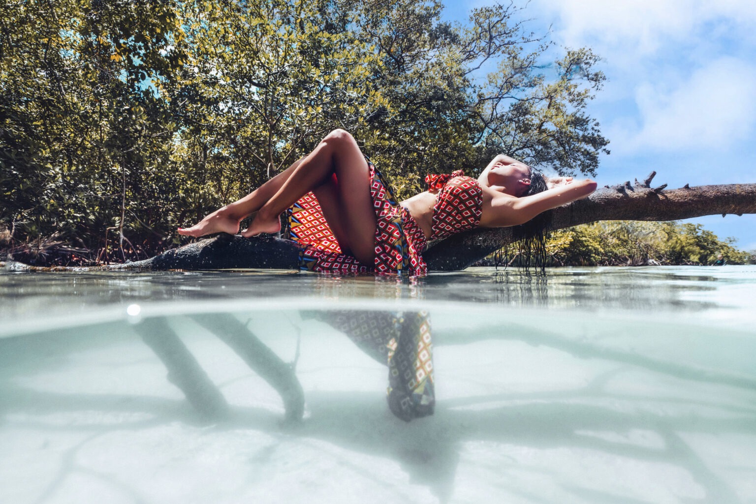 Editorial fashion photograph of a woman reclining gracefully on a mangrove branch in shallow turquoise water, wearing a red geometric-patterned two-piece set, captured by David Scarola Photography for Coastal Girls Co. on Jupiter Island.David Scarola Photography, Coastal Girls Co., Jupiter Island fashion photography, Palm Beach fashion photographer, fine art editorial, resort fashion campaign, luxury lifestyle photography, underwater fashion art, Florida coastal editorial, high-end boutique photoshoot, Jupiter Island elegance, Palm Beach photographer, artistic fashion portraiture.