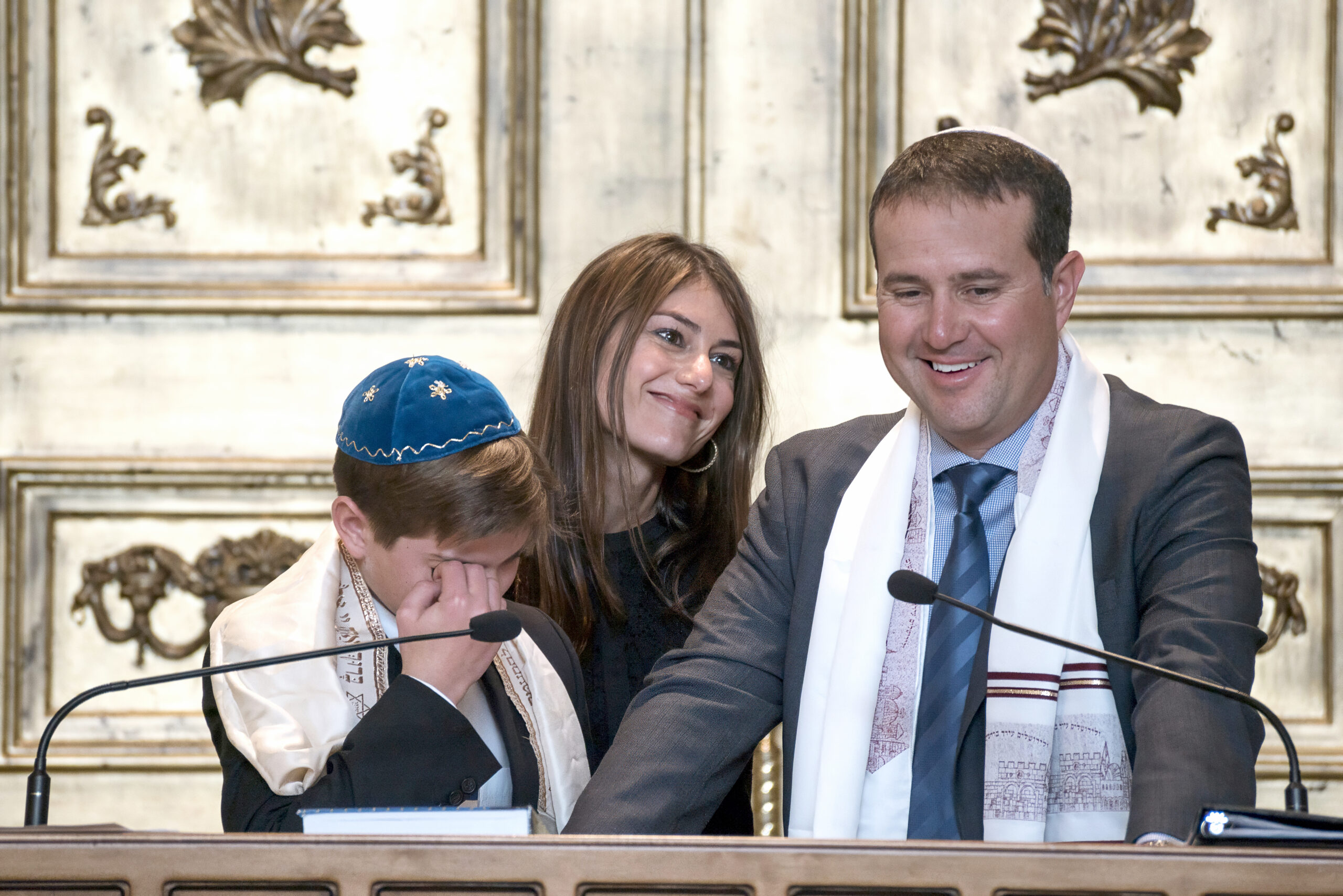 Emotional moment during Matthew Smith’s Bar Mitzvah at Temple Beth Am in Jupiter, Florida. The young man stands beside his smiling father and proud sister, wiping away tears of joy at the bimah as the family shares a deeply meaningful milestone in the sacred space.