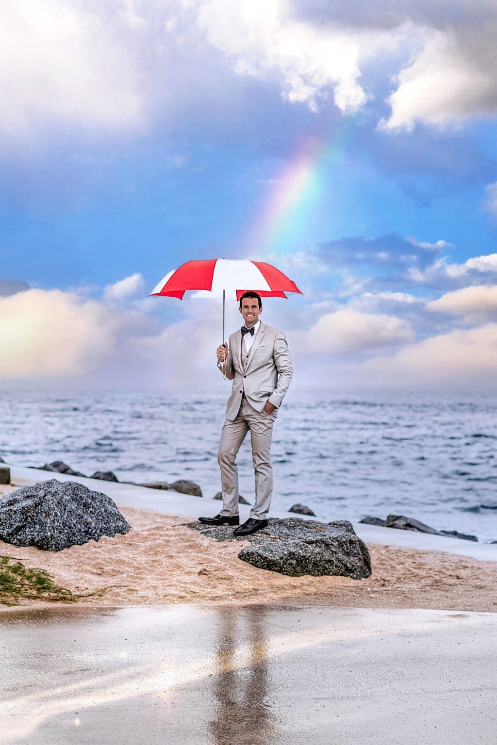 Groom in a light suit holding a red and white umbrella under a glowing rainbow on Palm Beach Island, captured by Florida’s best wedding photographer, David Scarola — a top-rated portrait artist and master of fine-art wedding photography by the sea.