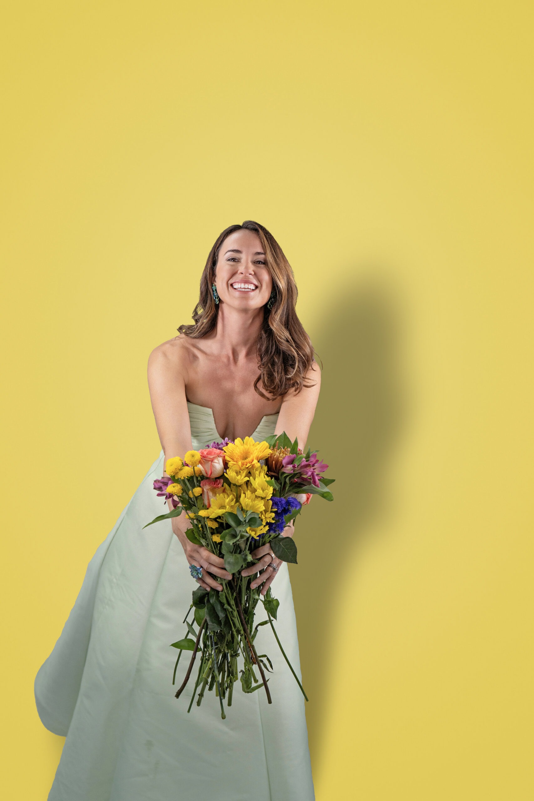 Singer-songwriter Britt Drozda photographed in studio, radiant and joyful, holding a vibrant bouquet of flowers against a bright yellow background. Captured by David Scarola Photography, this creative portrait embodies the color, confidence, and charisma of a modern female artist.