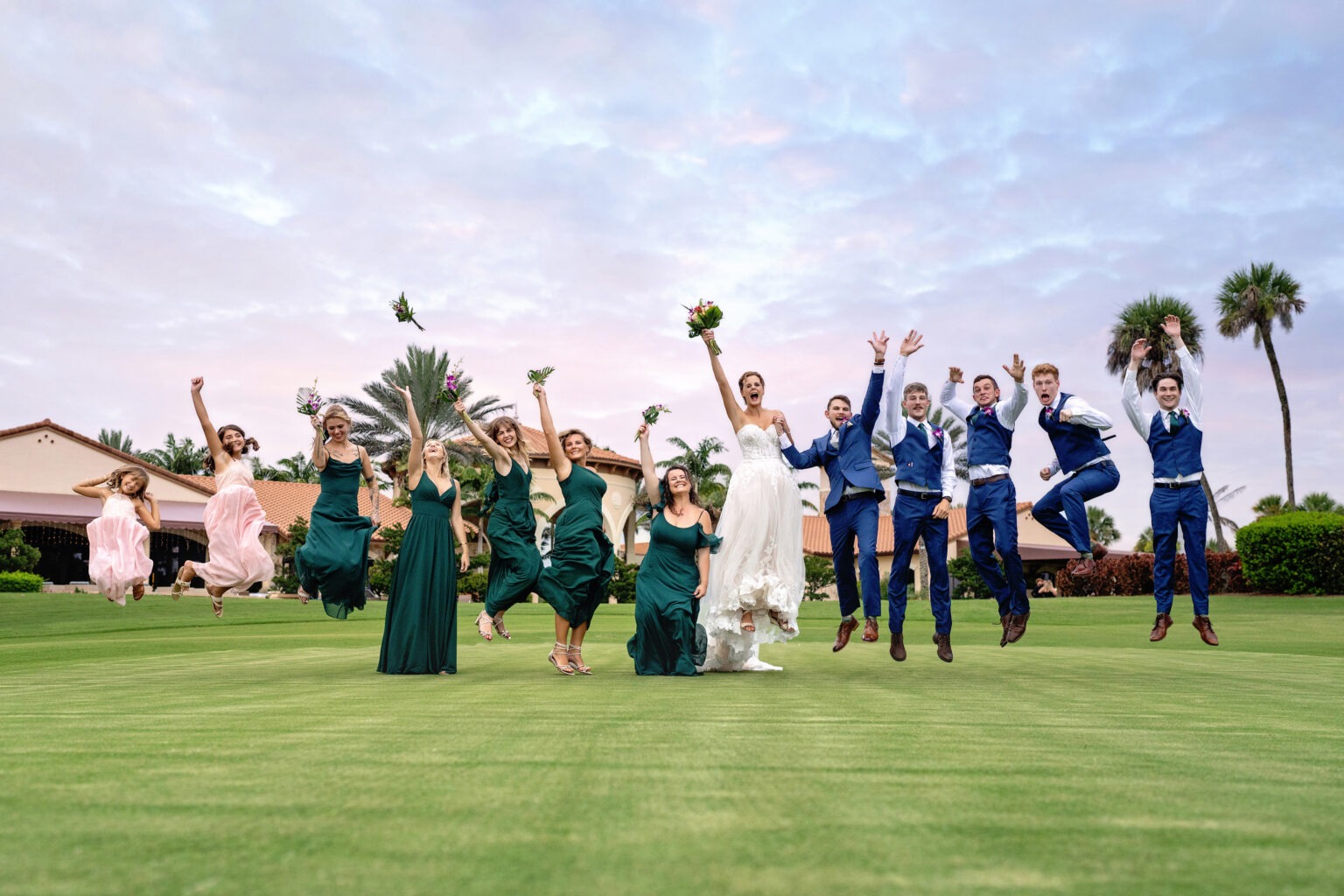 Joyful wedding party midair during a fun jump shot on the golf course at Jupiter Country Club — bridesmaids in green and pink, groomsmen in navy, and the bride leading the celebration — captured in vibrant fine-art style by David Scarola, Florida’s best wedding photographer and top-rated portrait artist specializing in dynamic, story-driven wedding imagery.
