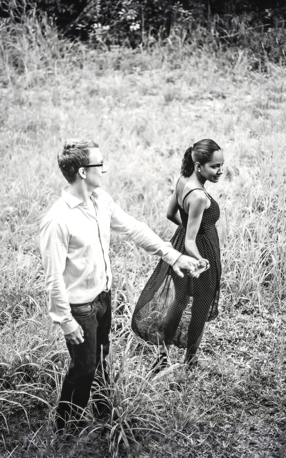 Engaged couple walking hand in hand through a sunlit coastal field by the beach on Jupiter Island, Florida — captured in timeless black and white. The bride-to-be’s dress flows softly in the breeze as their connection radiates through the frame, photographed with emotion and artistry by David Scarola, Florida’s best engagement and fine-art wedding photographer renowned for cinematic storytelling and soulful portraiture.
