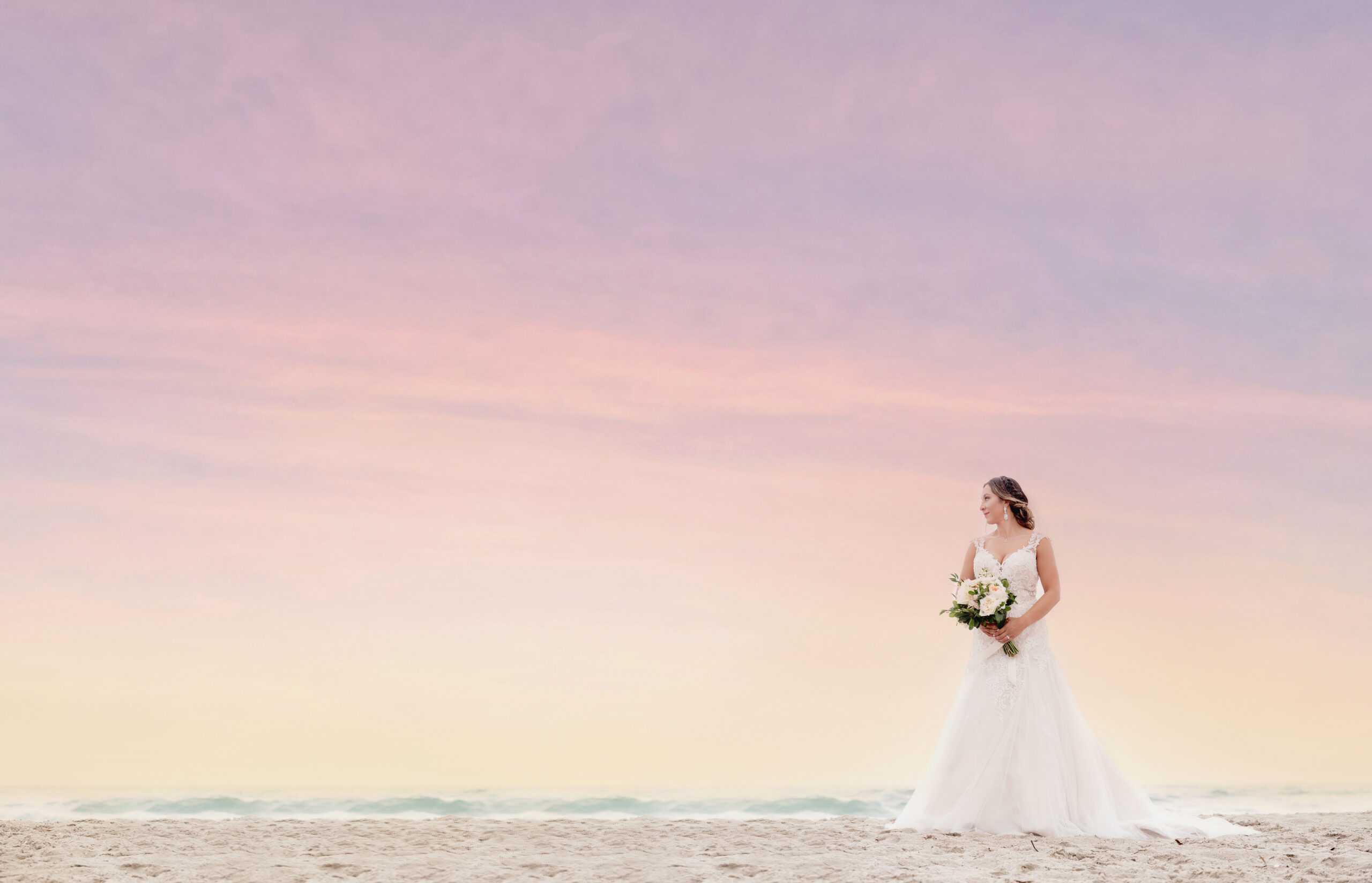 Bride standing gracefully on the sandy beach at sunset in Palm Beach, Florida, surrounded by pastel skies of pink and gold — captured in fine-art elegance by David Scarola, Florida’s best wedding photographer and master portrait artist known for luminous, ocean-inspired wedding imagery.
