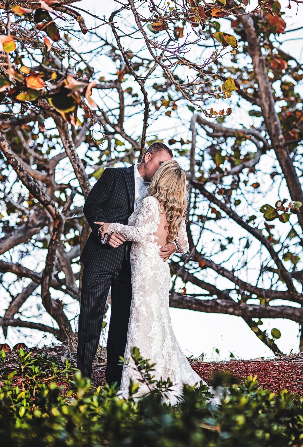 Bride and groom share a romantic kiss under sea grape trees at Jupiter Beach Resort on Jupiter Island, Florida. Captured by David Scarola Photography, this fine-art wedding portrait embodies love, elegance, and coastal serenity with cinematic lighting and natural emotion.