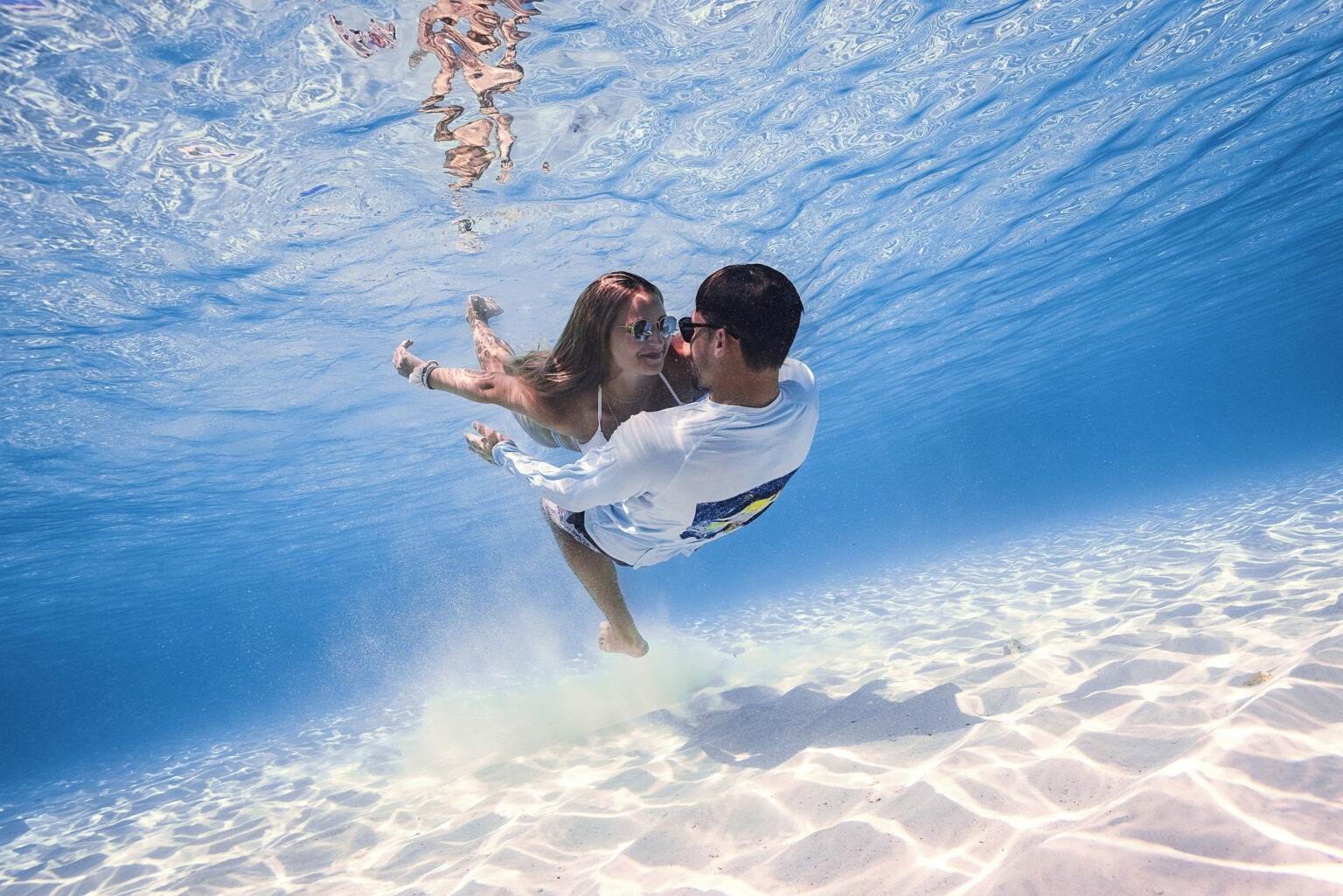 Underwater editorial photograph of a couple embracing gracefully in crystal-clear turquoise water, sunlight rippling across the ocean floor, captured by David Scarola Photography for Coastal Girls Co. and Jupiter Magazine’s Ocean Magic fashion and art campaign.David Scarola Photography, Coastal Girls Co., Jupiter Magazine, Ocean Magic campaign, underwater photography, fine art photographer Florida, Jupiter Island editorial, Palm Beach fashion photographer, luxury lifestyle photography, ocean editorial art, underwater couple portrait, resort campaign photography, South Florida fashion photography, professional underwater portraits, best underwater photographer in Florida.