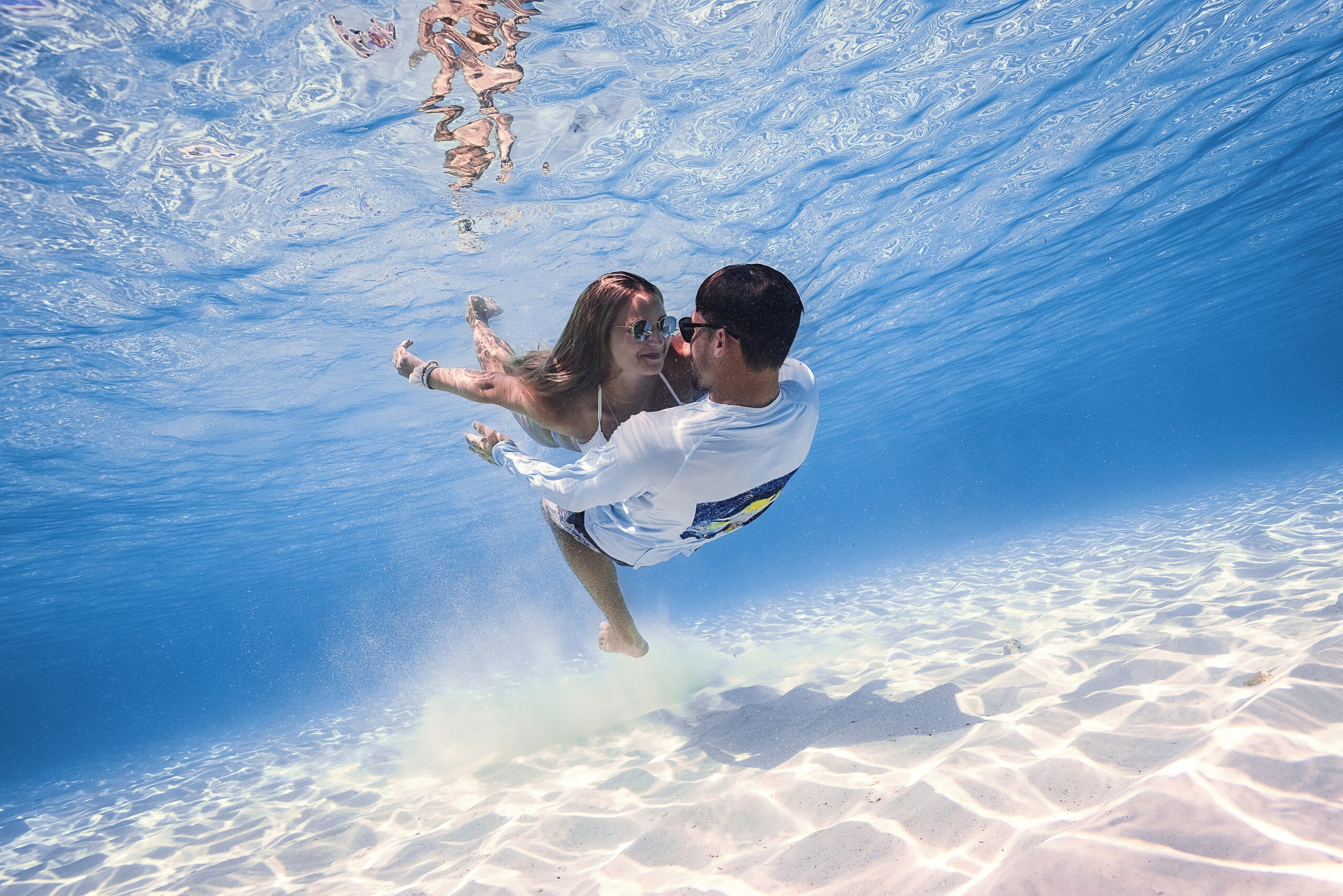 Underwater editorial photograph of a couple embracing gracefully in crystal-clear turquoise water, sunlight rippling across the ocean floor, captured by David Scarola Photography for Coastal Girls Co. and Jupiter Magazine’s Ocean Magic fashion and art campaign.David Scarola Photography, Coastal Girls Co., Jupiter Magazine, Ocean Magic campaign, underwater photography, fine art photographer Florida, Jupiter Island editorial, Palm Beach fashion photographer, luxury lifestyle photography, ocean editorial art, underwater couple portrait, resort campaign photography, South Florida fashion photography, professional underwater portraits, best underwater photographer in Florida.