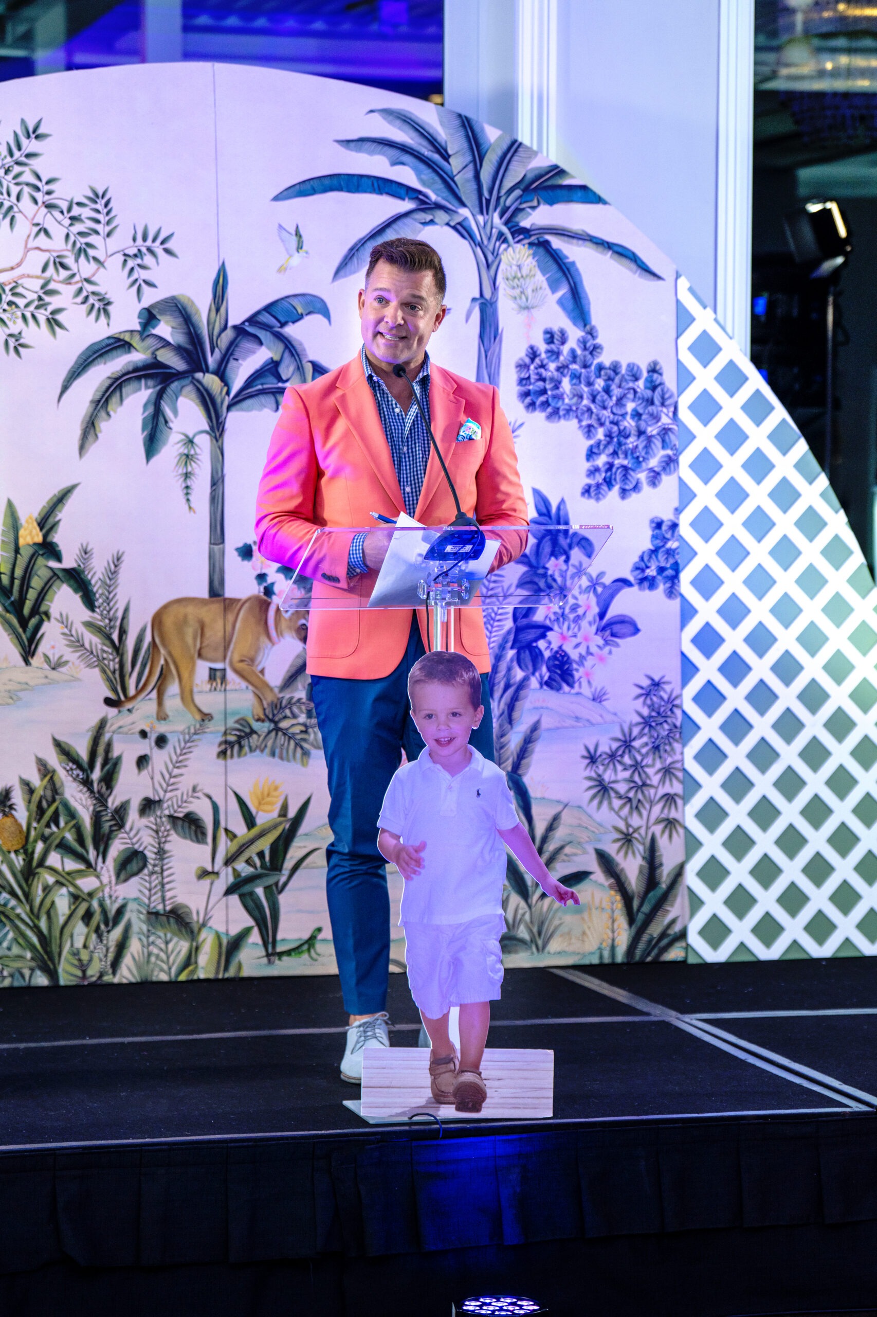 Jay Cashmere, event emcee, speaks on stage at the Live Like Jake Gala at The Pelican Club in Jupiter Island, Florida. Dressed in a coral blazer, he stands beside a display of young Jake’s photo, honoring the foundation’s life-saving mission.Jay Cashmere emcee, Live Like Jake Gala, Pelican Club Jupiter Island, charity gala photography, luxury event photographer Florida, Palm Beach gala photographer, philanthropic event photography, David Scarola Photography, global event coverage, high-end gala photography, drowning prevention charity, Jupiter Island event photographer, professional event emcee USA, Palm Beach luxury events.