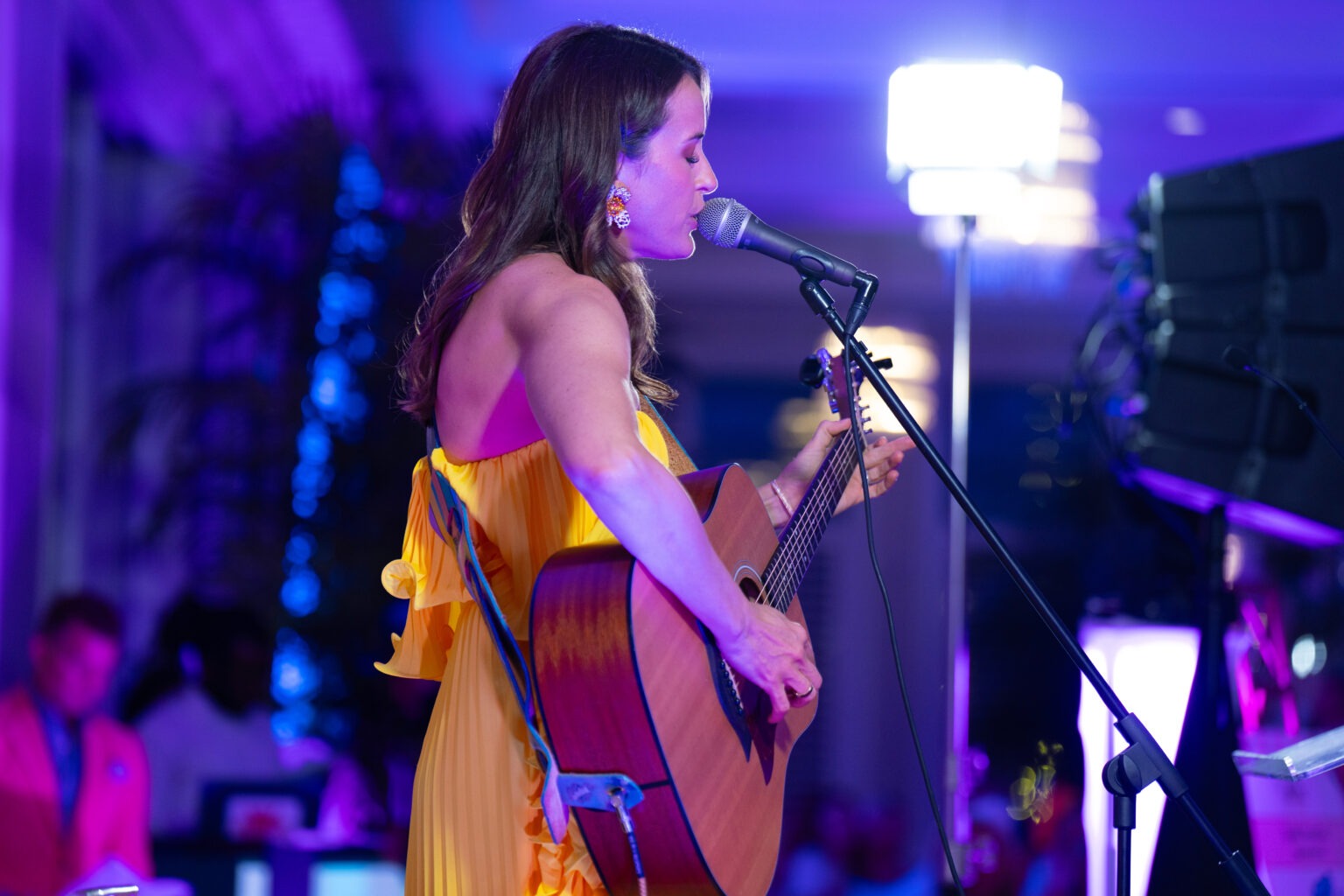 Female singer performing live at the Live Like Jake Gala at The Pelican Club in Jupiter Island, Florida. She’s wearing a flowing yellow dress and playing an acoustic guitar under vibrant purple and blue lighting, with the audience softly visible in the background.Live Like Jake Gala, Britt Drozda performance, Pelican Club Jupiter Island, Jupiter Island event photographer, Palm Beach gala photography, Florida charity event, corporate event photography, luxury event photography, David Scarola Photography, worldwide event coverage, nonprofit gala photographer, high-end event photographer.