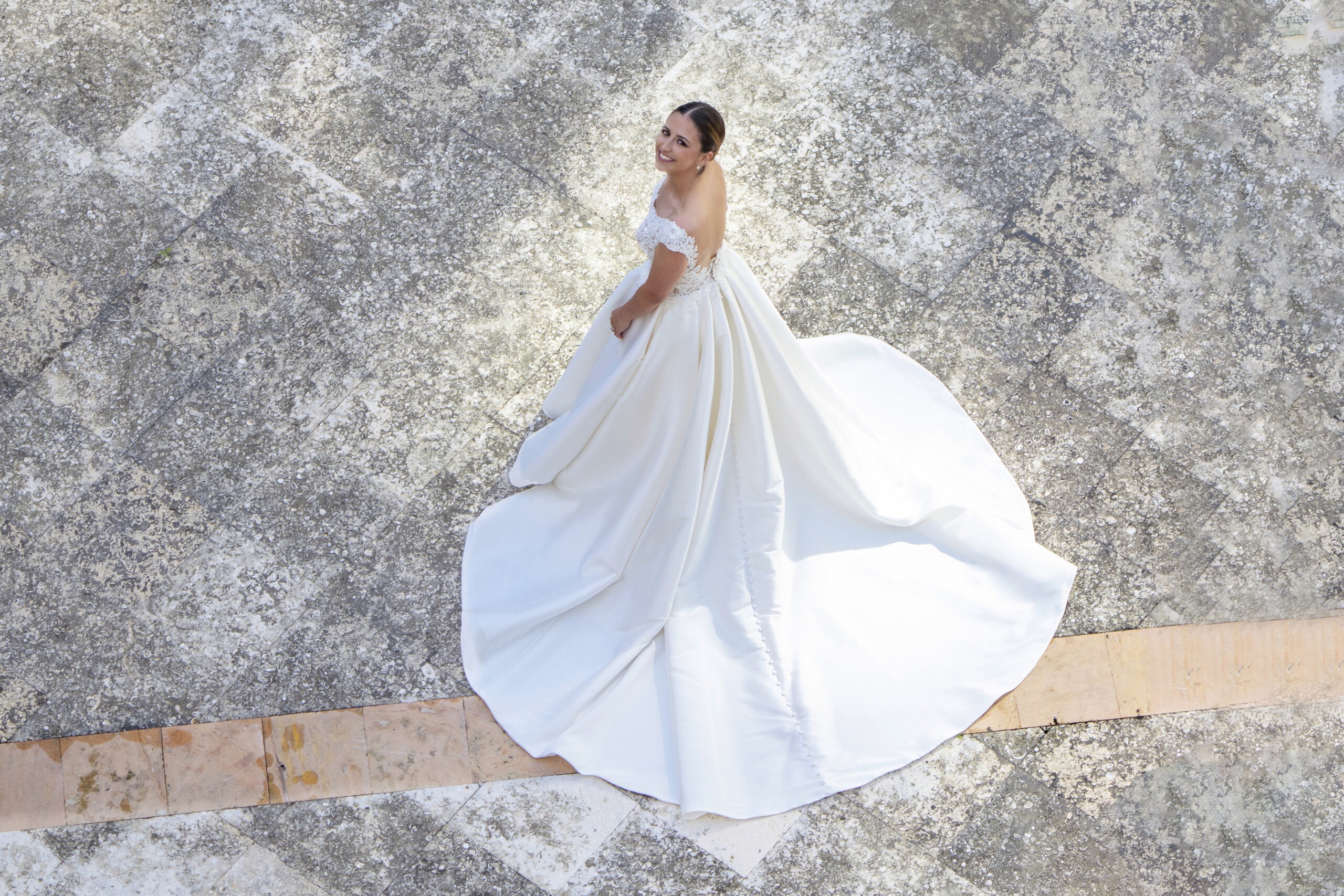 Bride in an elegant off-the-shoulder gown with a flowing train, smiling radiantly while standing on the textured stone courtyard of her Jupiter Island wedding — captured from above in soft natural light by David Scarola, Florida’s best wedding photographer and world-class fine-art portrait artist renowned for luxury coastal weddings and timeless elegance.