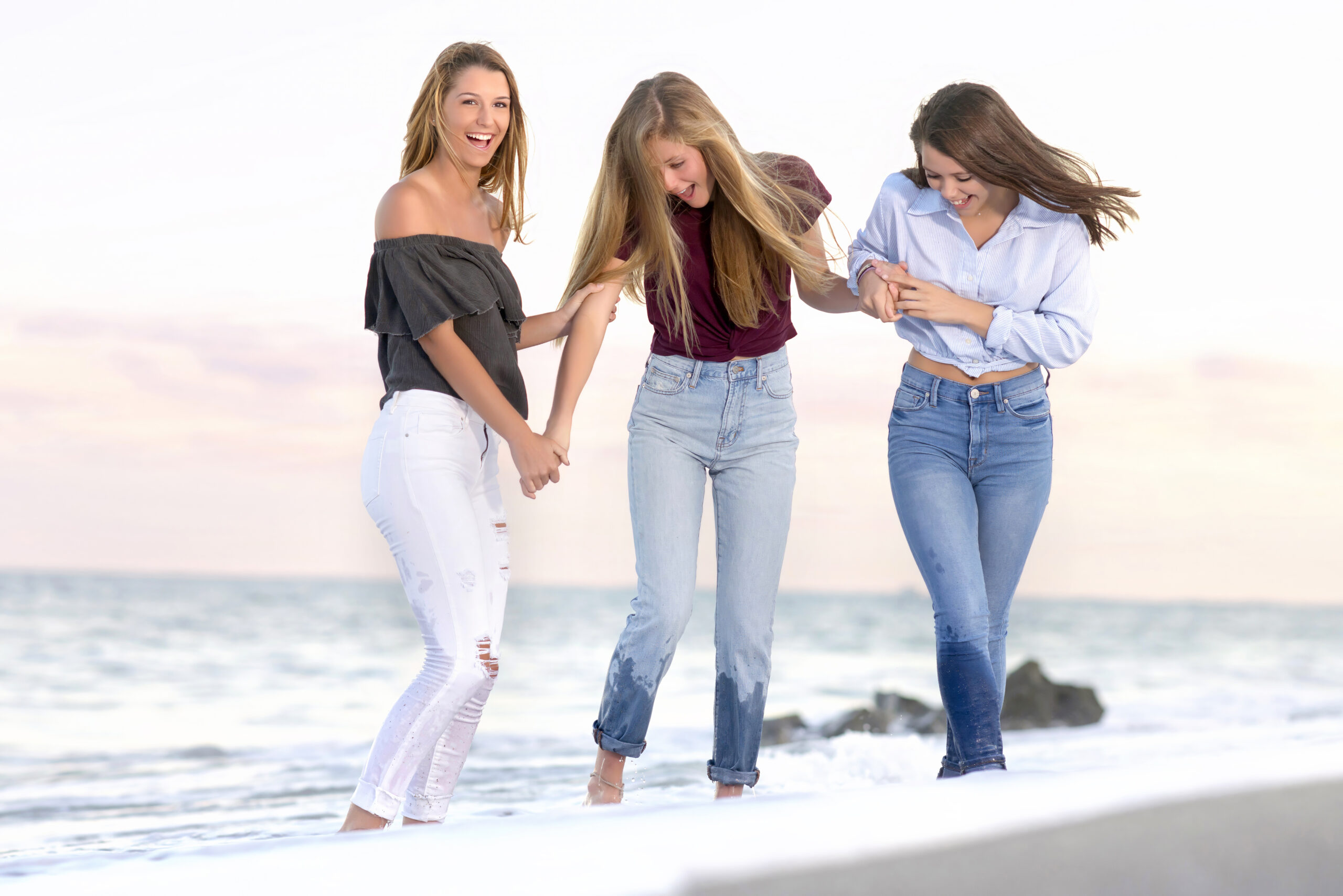 3 darling teenage sisters in the soft breaking surf at the beach on Palm Beach Island playing and laughing during their family photography session, a shoot with their parents, and cousins, with portrait artist David Scarola, both a master of the golden hour photo shoots, and one considered by many to be the best photographer in Palm Beach.