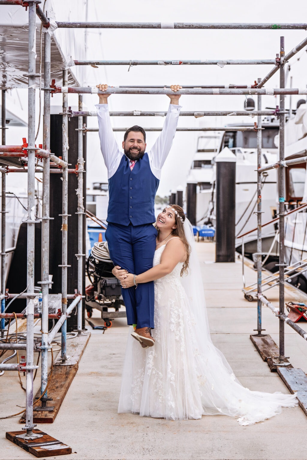Bride and groom sharing a playful wedding moment at Rybovich Marina in Palm Beach, with the groom hanging from scaffolding and the bride laughing and holding him up — photographed by David Scarola Photography.