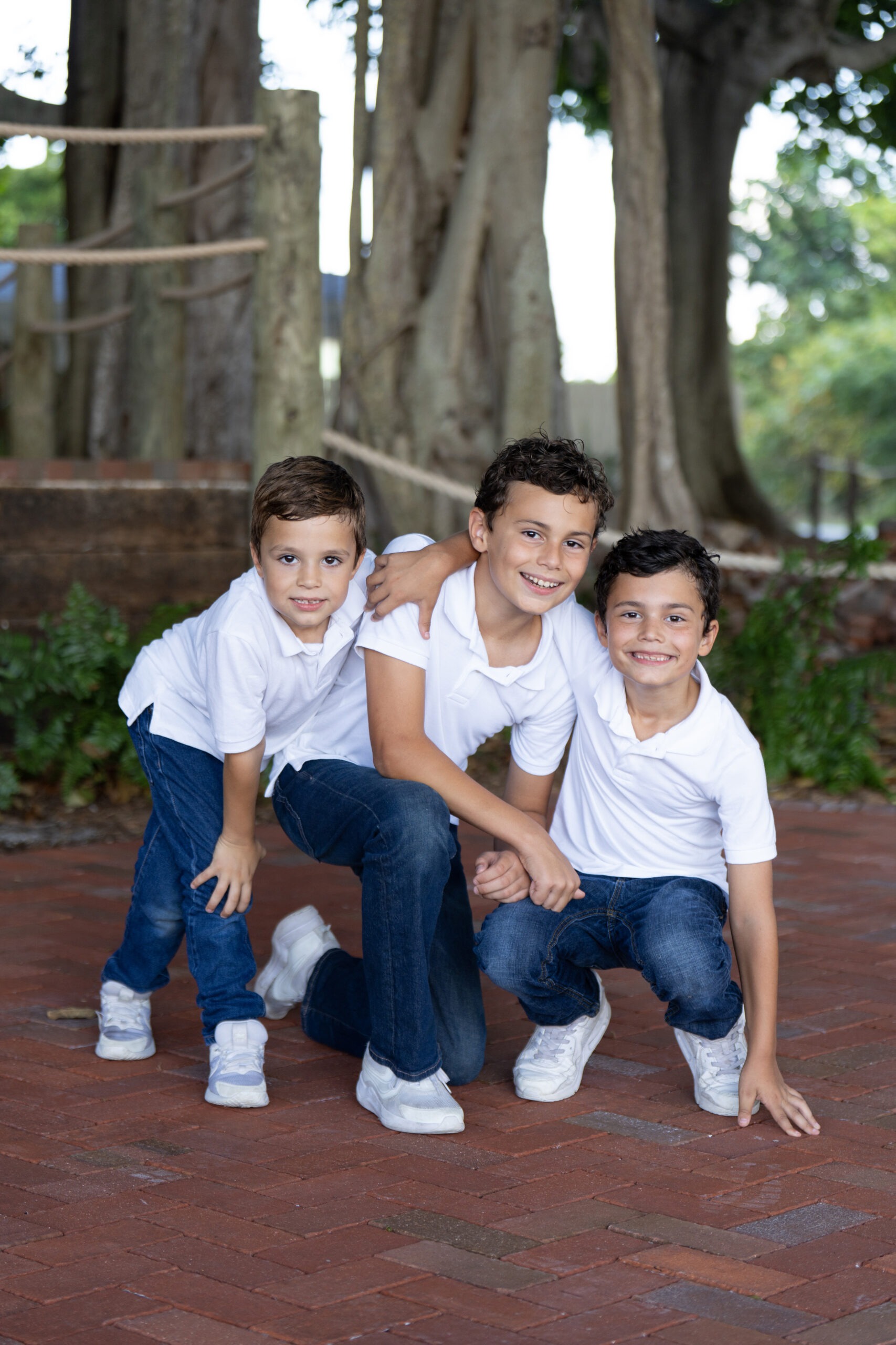 Three young brothers wearing white shirts and blue jeans smile and kneel together on a red-brick pathway beneath the towering banyan trees at the Jupiter Lighthouse Center in Florida.family photographer in Florida, best family portraits Jupiter Lighthouse, top portrait photographer Palm Beach, Jupiter Island family photography, Scarola Gallery portraits, Palm Beach County family photos, fine art family photography, master portrait artist Florida, Jupiter FL professional photographer, children and sibling photography Palm Beach.A heartwarming family portrait of three brothers captured at the Jupiter Lighthouse Center. Each child’s joy and bond is illuminated through the lens of master portrait artist David Scarola — Florida’s premier family photographer. Known for blending natural light, authentic emotion, and refined artistry, David’s portraits are timeless keepsakes of love and connection.