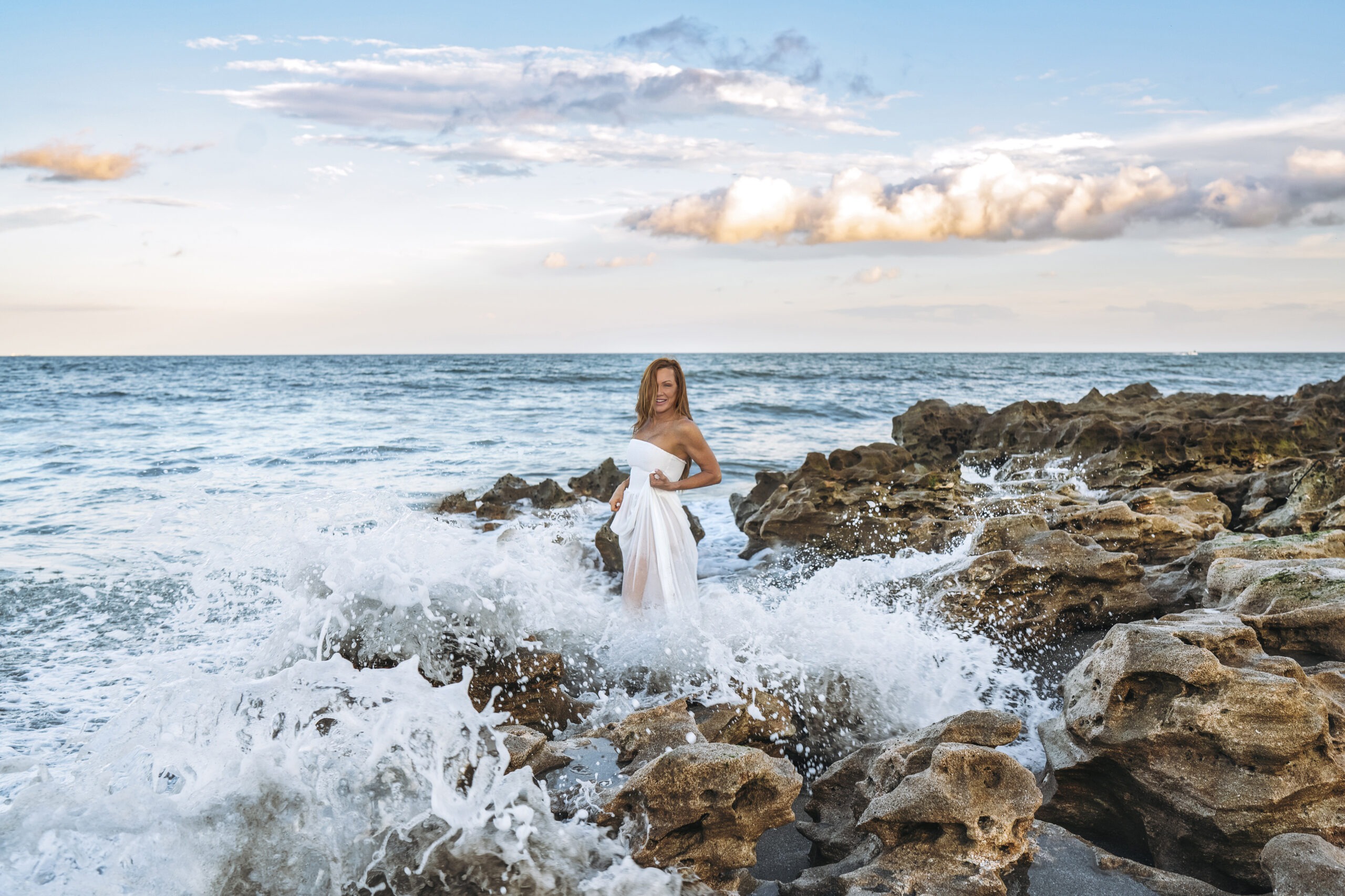Jupiter Island bride in a flowing white gown standing on the rugged coral rocks as ocean waves crash around her—epic “trash the dress” portrait captured in luminous coastal light by David Scarola, Florida’s best wedding photographer and master fine-art portrait artist.