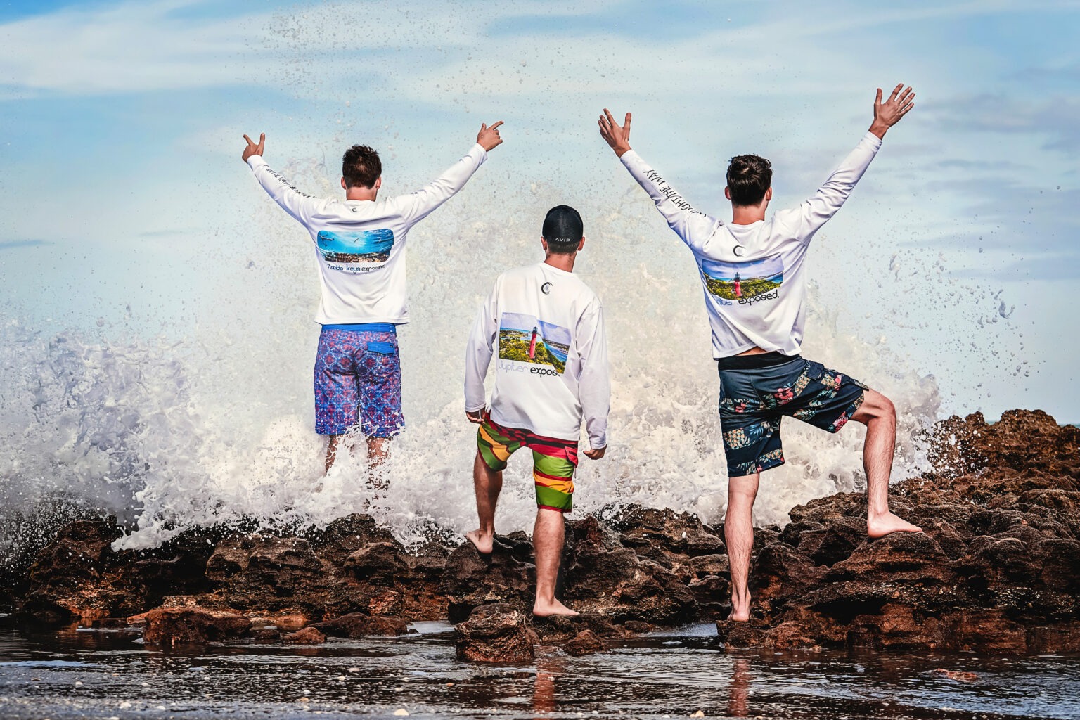 Three men stand triumphantly on the coral rocks at Coral Cove Beach in Jupiter Island, Florida, as ocean waves crash around them. They are wearing Exposure Solgear activewear sunshirts from the Jupiter.Exposed collection, featuring vibrant photographic prints of Jupiter Lighthouse and the Florida coastline. Captured by David Scarola Photography, showcasing lifestyle, fashion, and the spirit of the Jupiter outdoors.Exposure Solgear, Jupiter.Exposed apparel, Jupiter Island photography, Coral Cove Beach, Jupiter Florida activewear, lifestyle photo shoot Florida, men’s sun protection shirts, fashion and surf wear, coastal lifestyle brand photography, David Scarola Photography, professional fashion photographer Florida, outdoor sportswear marketing campaign.