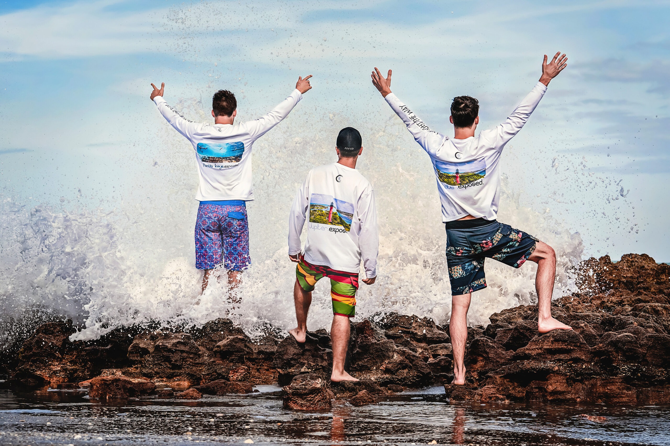 Three men stand triumphantly on the coral rocks at Coral Cove Beach in Jupiter Island, Florida, as ocean waves crash around them. They are wearing Exposure Solgear activewear sunshirts from the Jupiter.Exposed collection, featuring vibrant photographic prints of Jupiter Lighthouse and the Florida coastline. Captured by David Scarola Photography, showcasing lifestyle, fashion, and the spirit of the Jupiter outdoors.Exposure Solgear, Jupiter.Exposed apparel, Jupiter Island photography, Coral Cove Beach, Jupiter Florida activewear, lifestyle photo shoot Florida, men’s sun protection shirts, fashion and surf wear, coastal lifestyle brand photography, David Scarola Photography, professional fashion photographer Florida, outdoor sportswear marketing campaign.