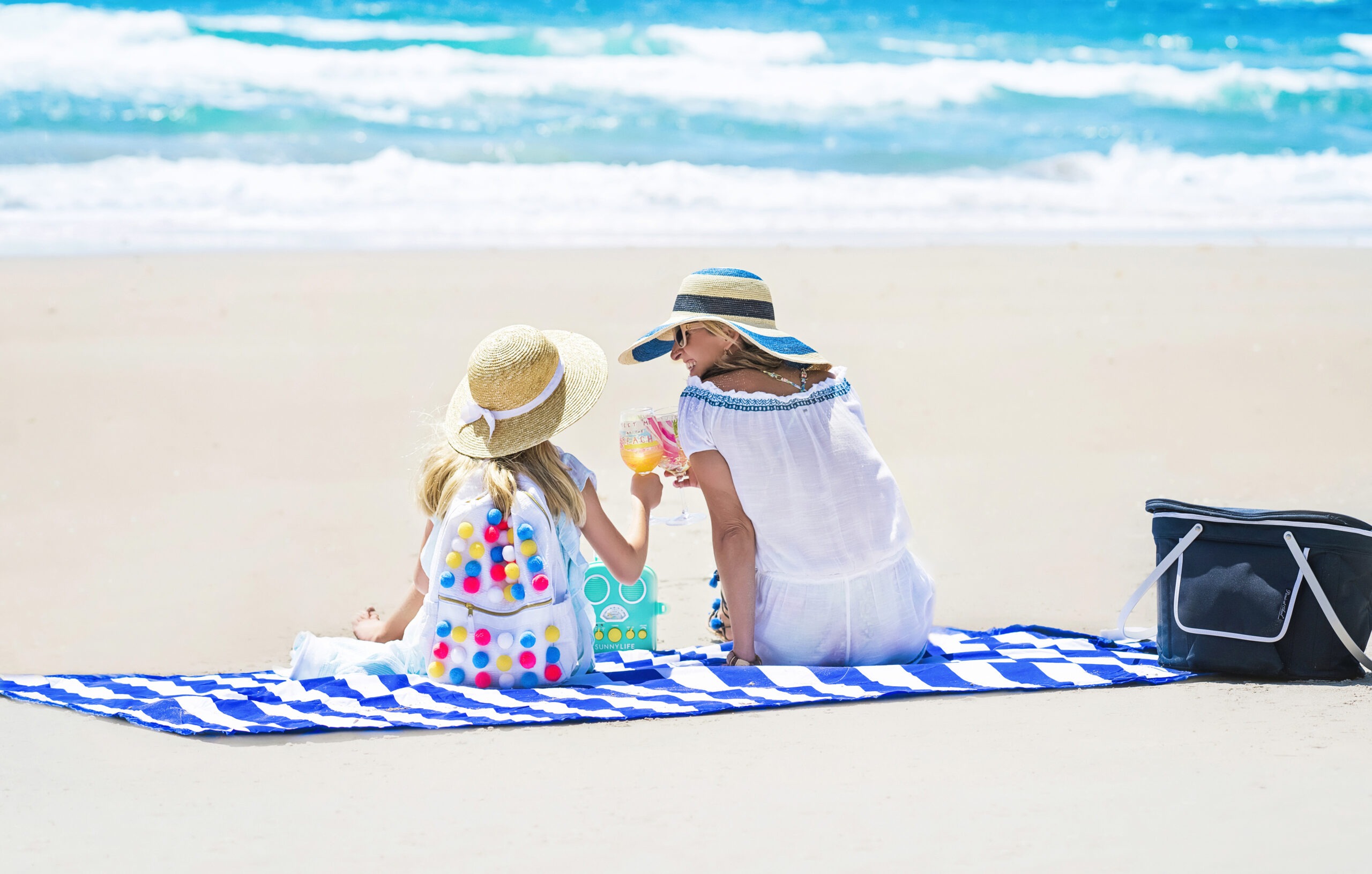 Mother and daughter enjoying a sunny day at the beach on Palm Beach Island, seated on a bold blue-and-white striped Palm Beach Crew blanket. The little girl wears a straw hat with a white ribbon and a colorful pom-pom backpack, while her mother, also in a wide-brimmed hat and white dress, smiles as they clink glasses of sparkling lemonade beside the turquoise Atlantic Ocean. Photographed by David Scarola for Nikiani Inc., showcasing Palm Beach Crew’s coastal lifestyle products that make moms’ lives easier.Nikiani Inc., Palm Beach Crew, Palm Beach photographer, Worth Avenue beach, mother-daughter lifestyle shoot, coastal fashion photography, beach products for moms, luxury family brand Florida, David Scarola Photography, professional commercial photographer Palm Beach, Jupiter Island resort photography.