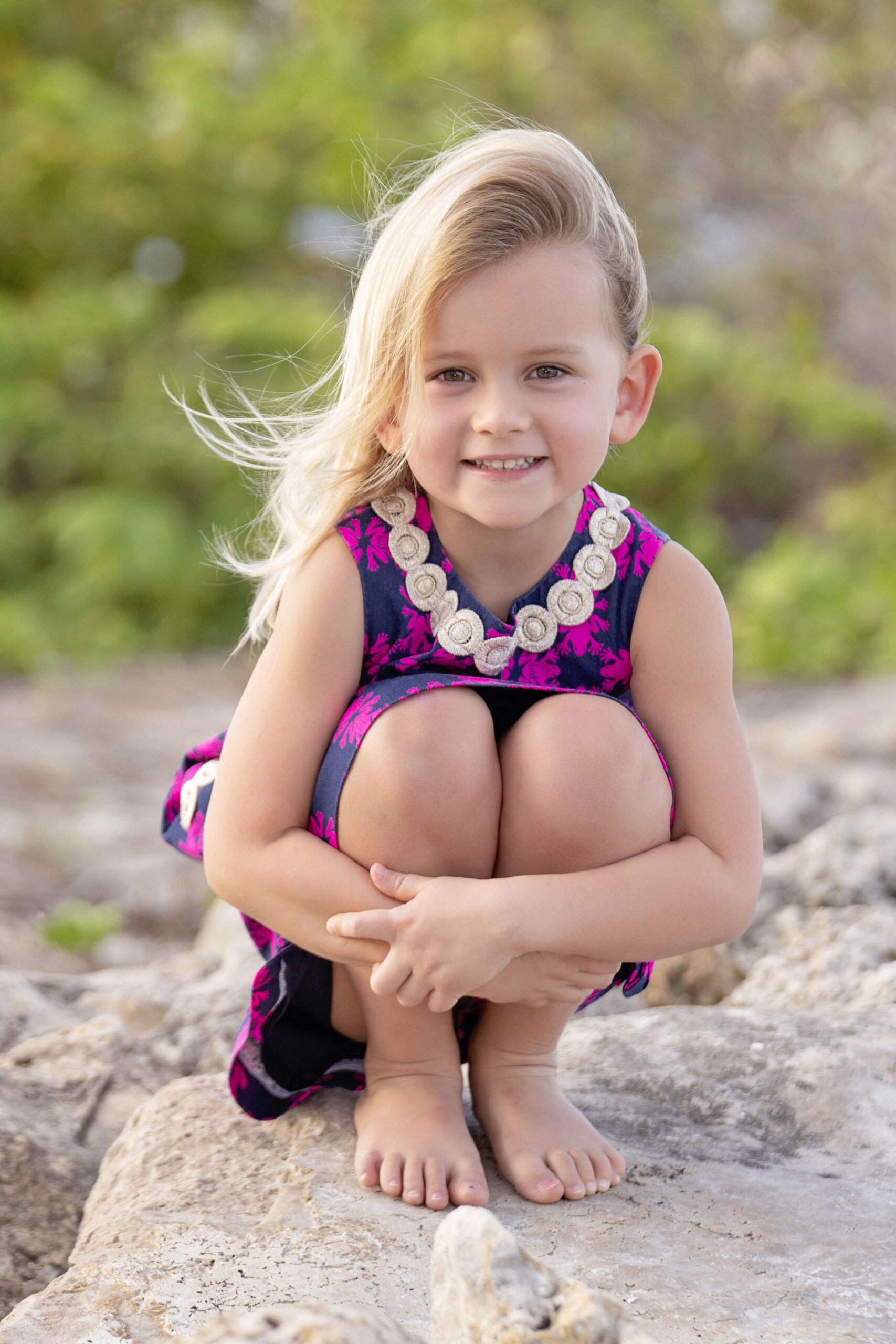 A young blonde girl crouches barefoot on the rocks at Dubois Park in Jupiter, Florida, wearing a navy and fuchsia floral dress with a decorative collar. Her hair catches the ocean breeze as she smiles sweetly toward the camera. Captured by master portrait artist David Scarola Photography, this image radiates warmth, innocence, and natural South Florida beauty.David Scarola Photography, Jupiter Island children photographer, Palm Beach family photographer, Dubois Park photo shoot, Jupiter Lighthouse portraits, South Florida lifestyle photography, fine art family photography, beach children portraits, Palm Beach County professional photographer, Jupiter Florida portraits, natural light family photos, child portrait photographer South Florida, outdoor children photo sessions, Jupiter Island photography sessions, best family photographer Palm Beach, Scarola Style photography, fine art children’s portraits Florida, coastal family photography, candid child portraits Jupiter Beach.