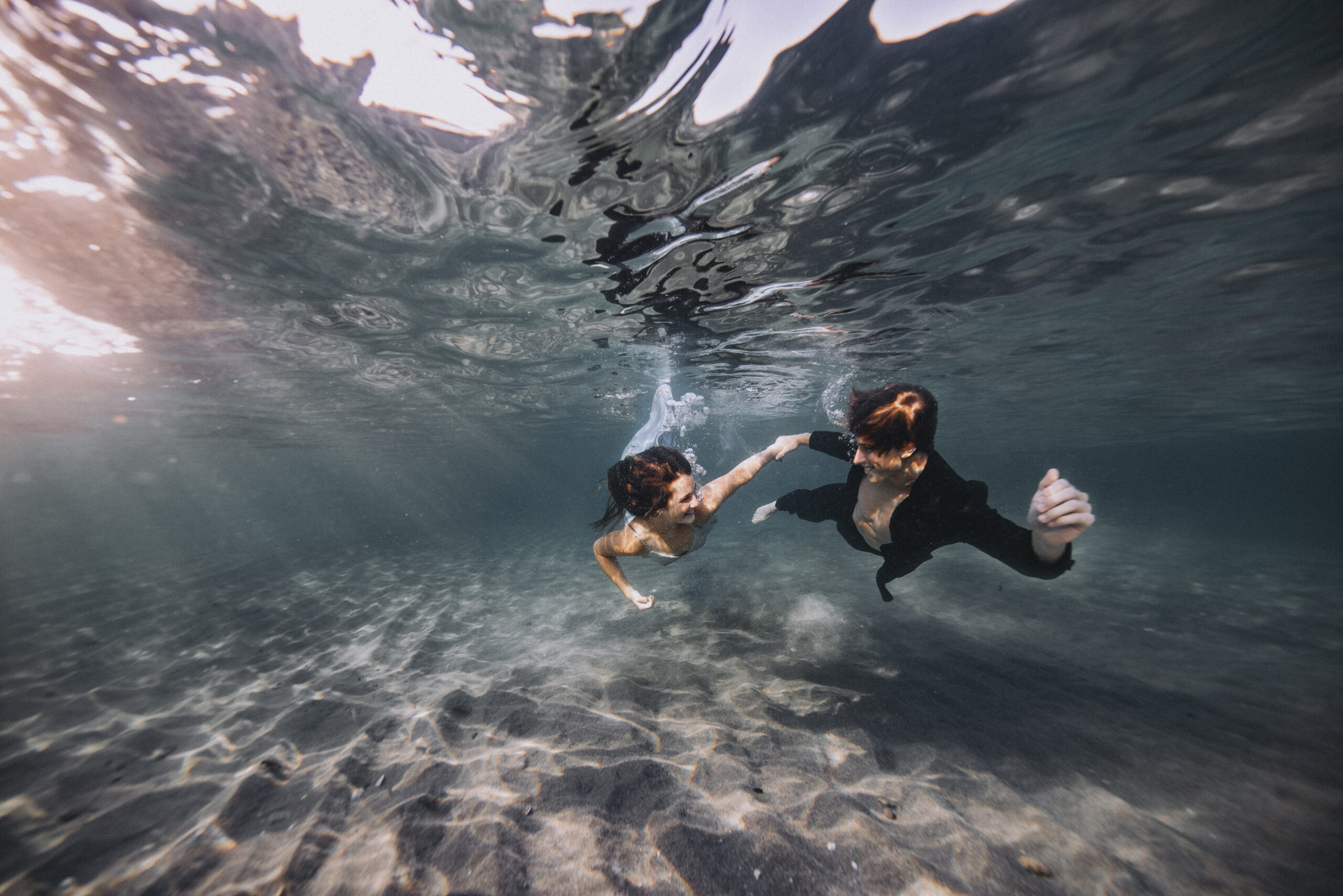 Underwater editorial photograph of a stylish couple gracefully swimming beneath the surface, light reflecting off rippled sand, captured in Palm Beach for a luxury fashion and art campaign by David Scarola Photography, inspired by Kirna Zabête and The Breakers Hotel.