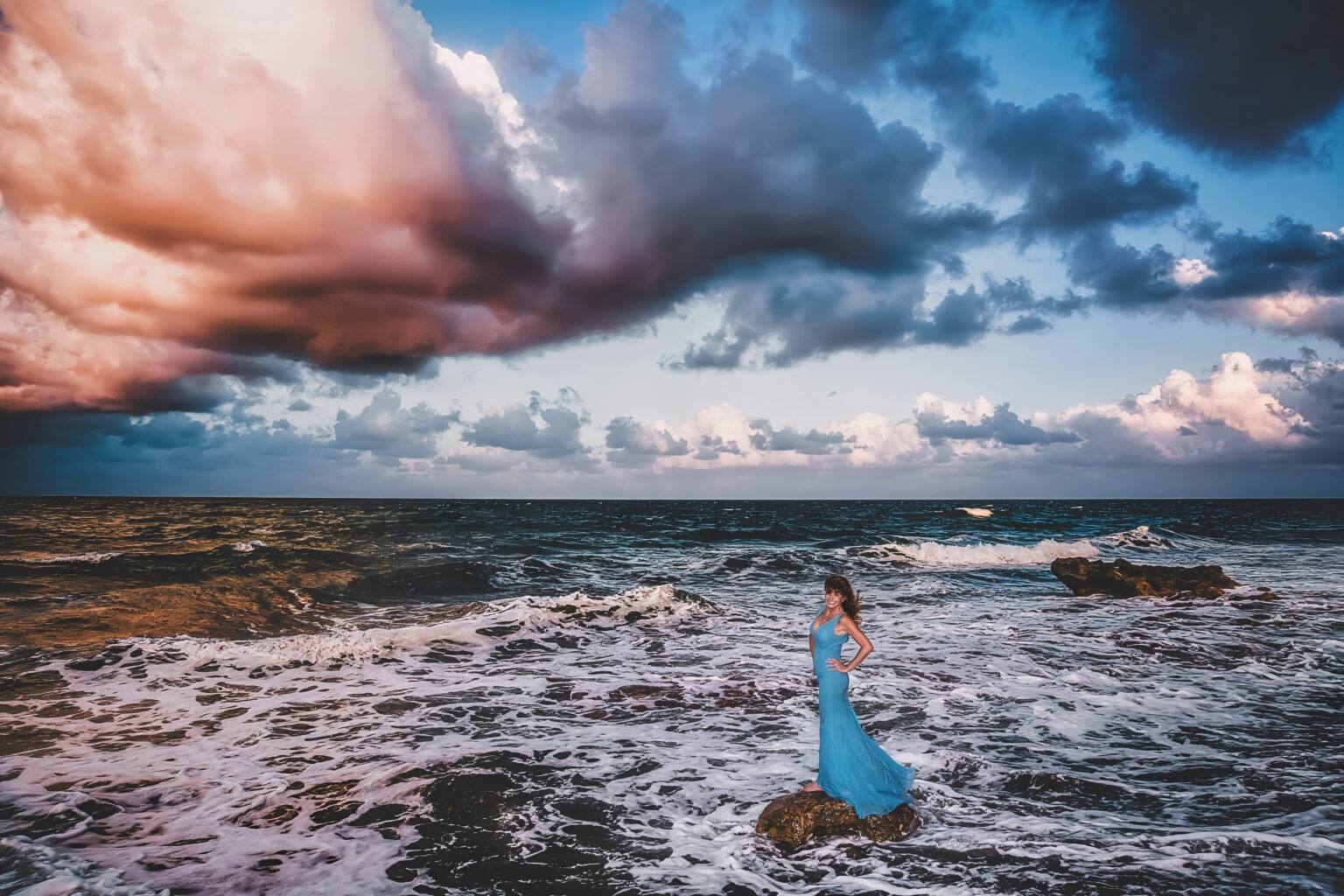 Fashion and lifestyle portrait of a woman standing confidently on a rock at Coral Cove Beach in Jupiter, Florida, photographed by David Scarola, one of the best fashion and lifestyle photographers in Palm Beach and the United States. She wears a flowing turquoise gown as waves crash around her, framed beneath a dramatic sky of pink and blue clouds. The image captures elegance, empowerment, and freedom through fine art fashion photography that merges beauty, power, and nature’s raw elements.