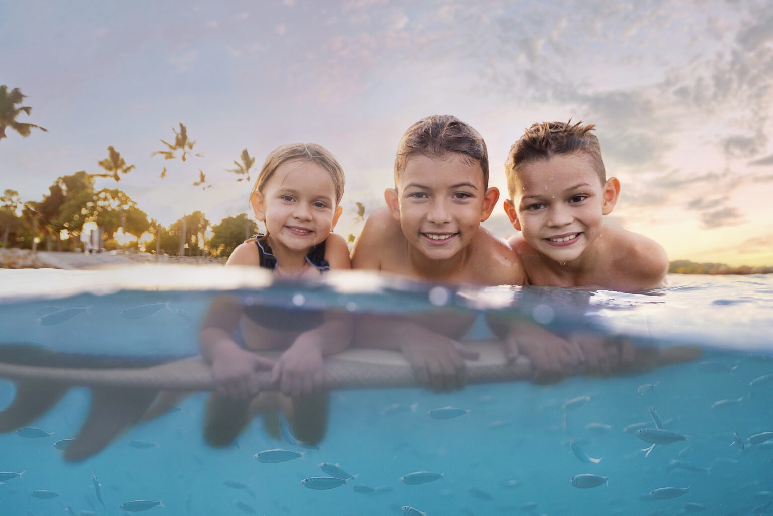 Three smiling children float together on a surfboard at sunset in crystal-clear blue water, captured half above and half below the surface at Dubois Park in Jupiter, Florida. A few small fish swim beneath them, while palm trees and warm evening light paint the background. Photographed by master portrait artist David Scarola, this image beautifully blends family joy, underwater artistry, and South Florida lifestyle.David Scarola Photography, Jupiter Island family photographer, underwater family photography Jupiter Florida, Palm Beach professional portrait artist, Dubois Park photo shoot, Jupiter Lighthouse photography, South Florida family portraits, underwater lifestyle photographer, fine art family photography, beach family photographer Palm Beach County, Jupiter Florida portrait sessions, master photographer David Scarola, family photo shoot at the beach, Palm Beach luxury family portraits, South Florida underwater portraits, editorial family photography, Jupiter Island photo artist, children lifestyle portraits Florida, underwater family art, best family photographer Palm Beach.