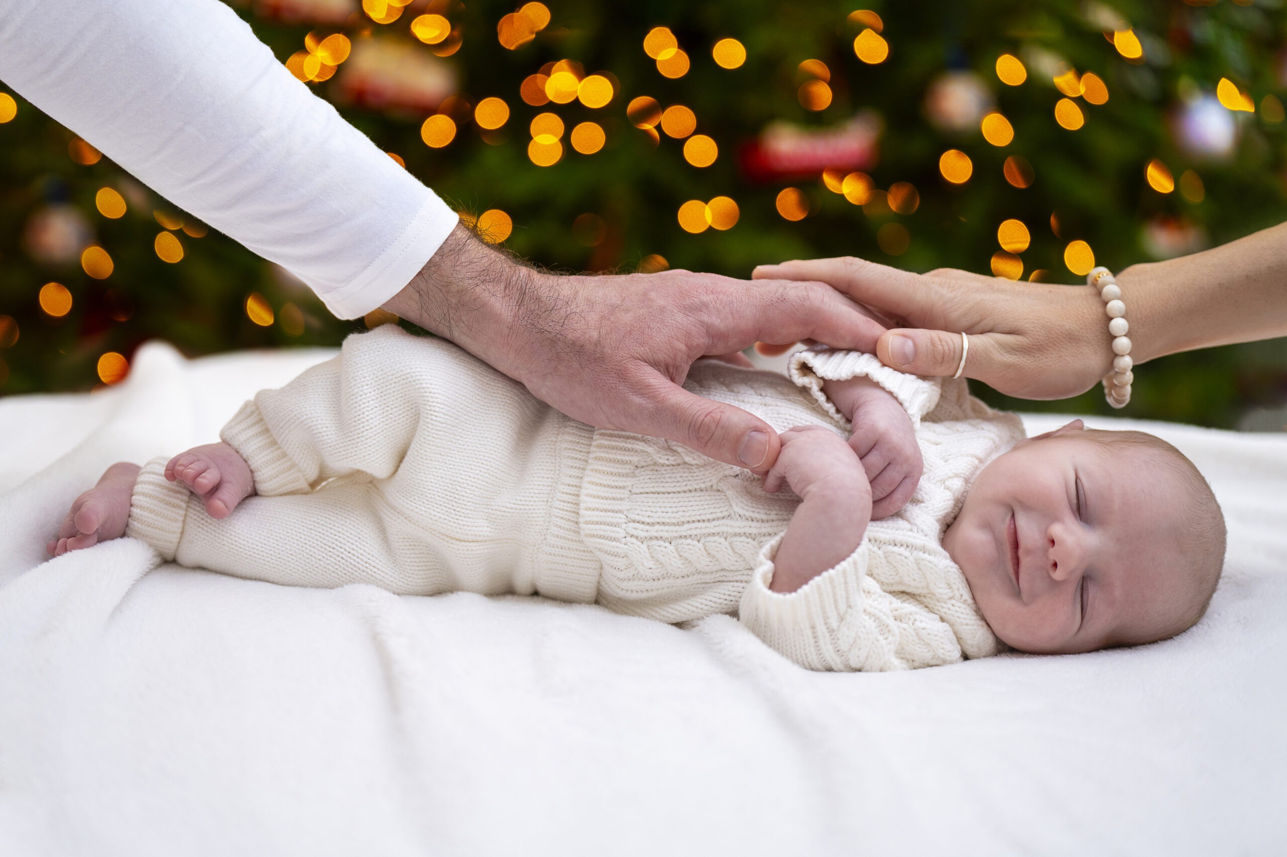 Smiling newborn baby named Vance during his first Christmas on Palm Beach Island, lying on a soft white blanket beneath glowing holiday lights. Parents’ hands gently touch his tiny fingers, symbolizing love, connection, and new life. Captured by David Scarola Photography, expert in Palm Beach newborn and family portrait photography.
