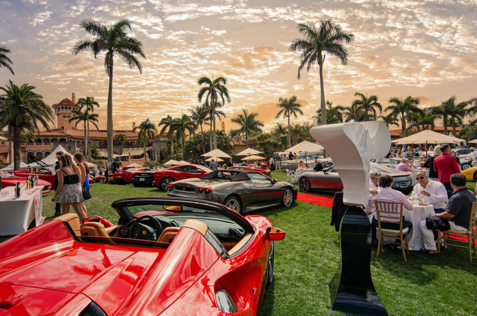 Luxury automotive showcase and social gathering on the lush lawn of Mar-a-Lago during The Palm Event. Exotic red Ferraris and other supercars gleam under a golden sunset surrounded by palm trees, art sculptures, and elegantly dressed guests enjoying fine dining outdoors.The Palm Event Palm Beach, Mar-a-Lago photography, luxury event photographer Florida, Palm Beach car show, exotic car photography, luxury lifestyle photography, corporate events Palm Beach, high end event photographer, worldwide event photography, fine art photographer, Ferrari Palm Beach, David Scarola Photography, elite events Florida.