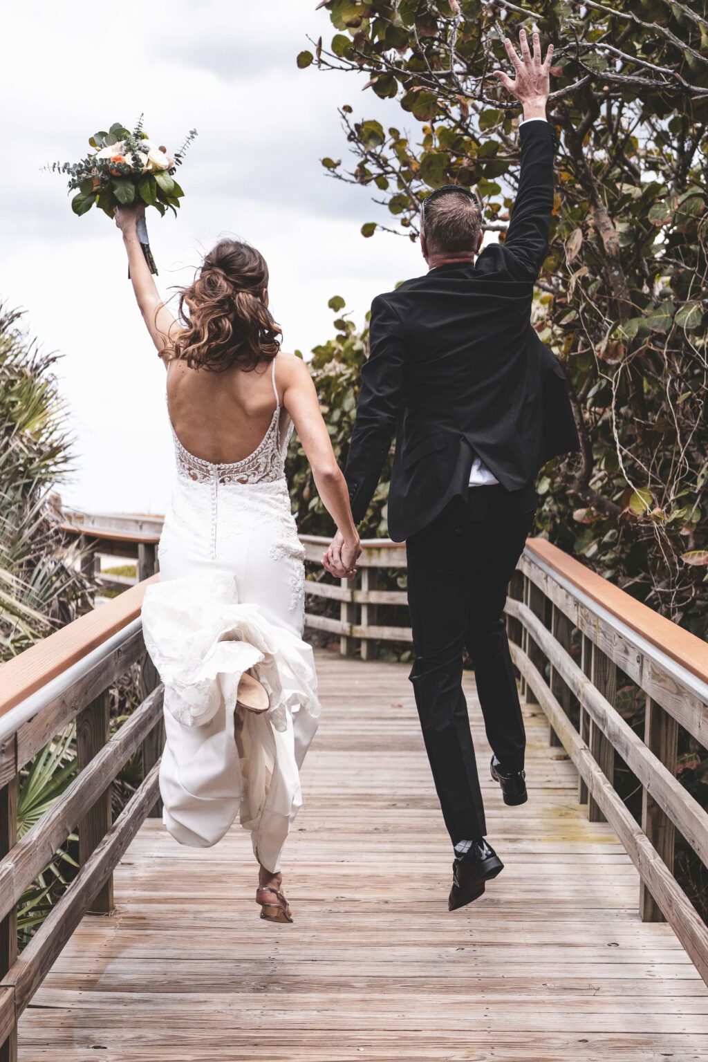 Bride and groom joyfully leaping on the wooden boardwalk at Coral Cove Park in Jupiter, Florida, moments after their beachside wedding ceremony. Captured by Palm Beach fine art photographer David Scarola, this vibrant image radiates post-ceremony bliss — the celebration of love, freedom, and light beneath Florida’s coastal sky.
