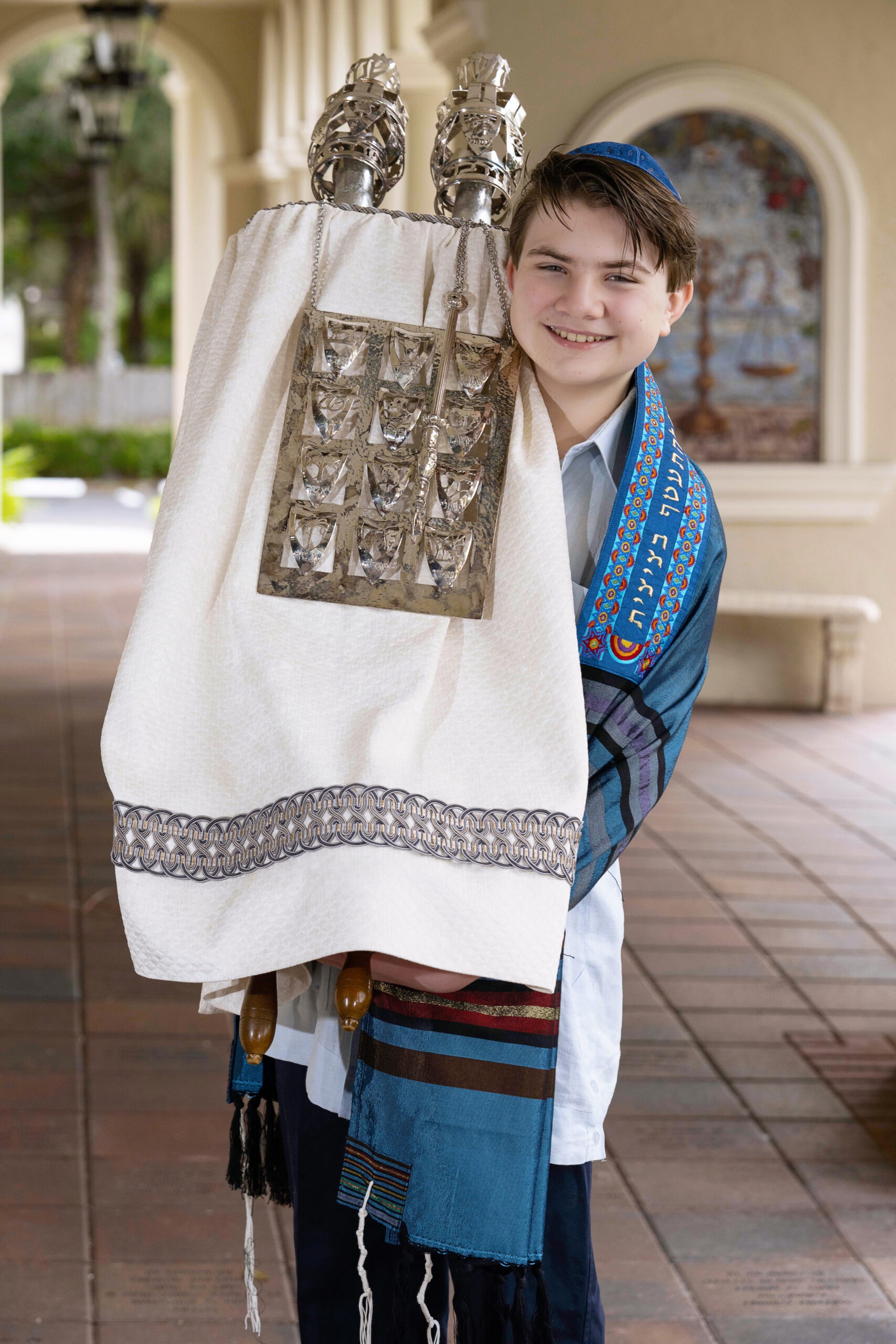 A young boy smiles proudly while holding the Torah during his Bar Mitzvah ceremony at Temple Beth David in Jupiter Island, wearing a blue tallit and kippah.Bar Mitzvah photography, Temple Beth David Jupiter Island, Jewish ceremony photographer, Palm Beach event photography, Torah portrait, David Scarola Photography, luxury family photographer, milestone celebration photography, high-end portrait artist, cultural event documentation, Jupiter Island photographer, professional event photographer, South Florida Bar Mitzvah photos.
