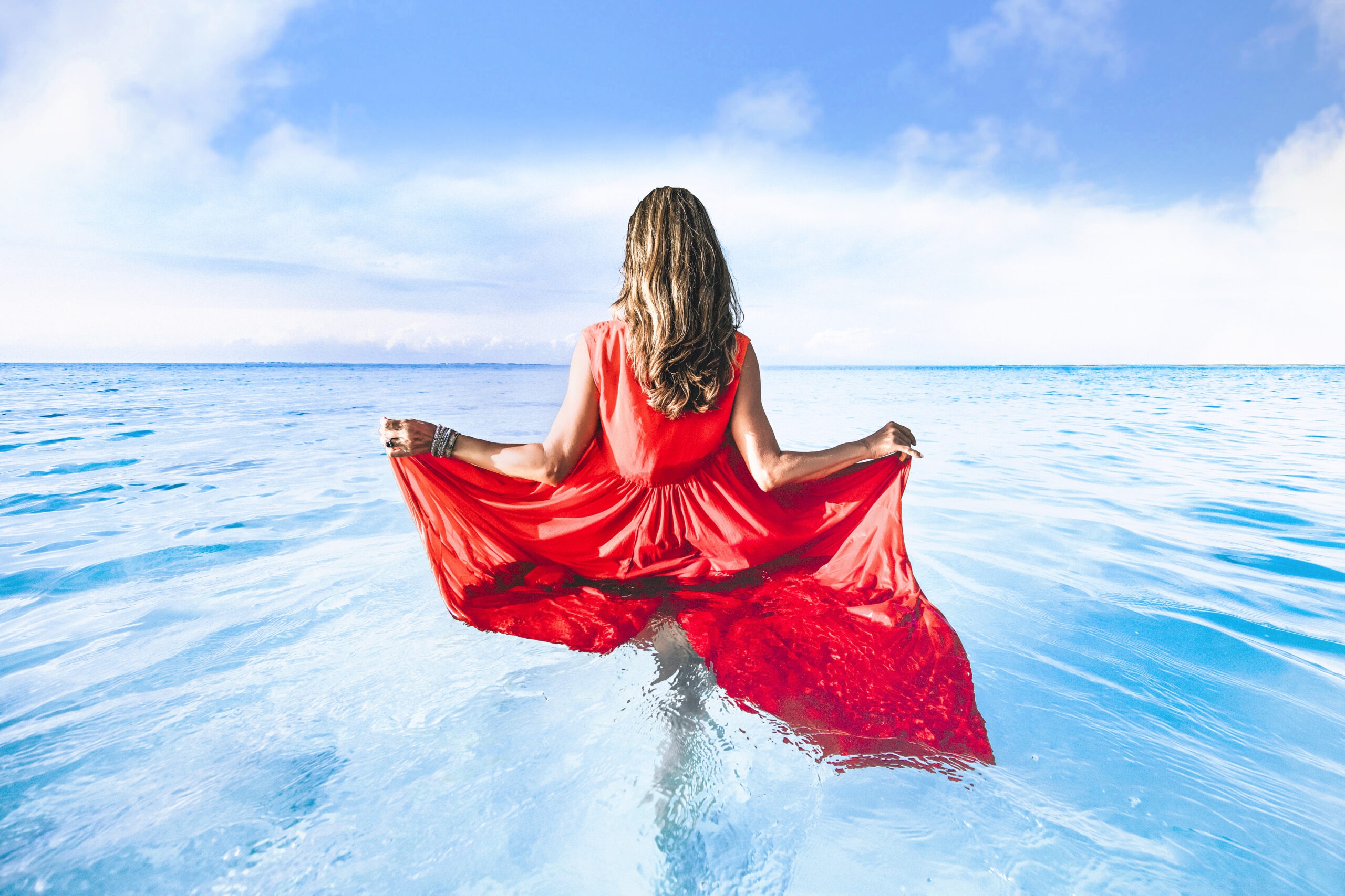 A woman in a flowing red dress stands waist-deep in the turquoise waters of Treasure Cay, Bahamas, holding the edges of her gown as sunlight reflects across the sea. The image captures the essence of high-end resort fashion and coastal elegance.each Boutique Sunset Harbour, Bahama Beach Resort photography, Treasure Cay fashion shoot, Abaco Islands catalogue, luxury resort fashion, Miami high-end boutique photography, David Scarola fine art photographer, Caribbean fashion photography, resortwear campaign Bahamas, professional fashion photographer Florida, destination editorial shoot, ocean lifestyle photography.