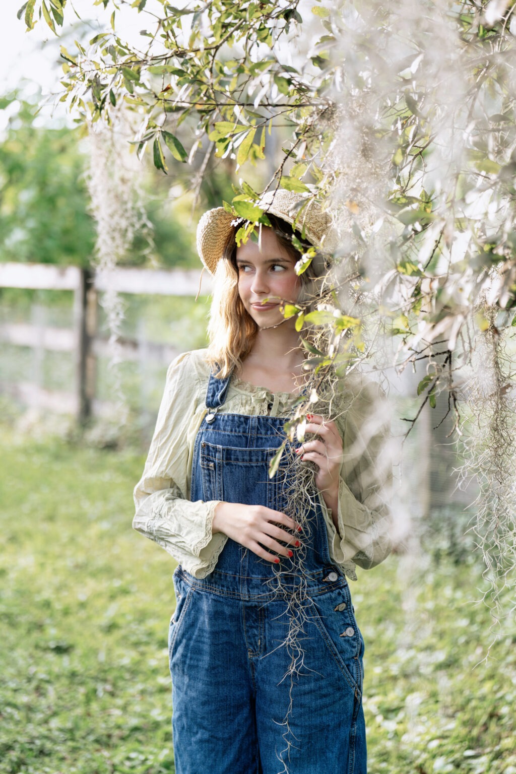 Senior portrait of Lucy in denim overalls and a straw hat, standing gracefully beneath Spanish moss in soft afternoon light at a Jupiter Island farm. Captured by David Scarola Photography, this image highlights natural beauty, confidence, and the authenticity of senior photography in Palm Beach County, Florida.senior portraits Jupiter Island, Palm Beach senior photographer, Jupiter Florida portrait photographer, outdoor senior photo ideas, natural light portrait photography, family photographer Palm Beach, David Scarola Photography, fine art portrait artist Florida, senior pictures South Florida, lifestyle senior photography.