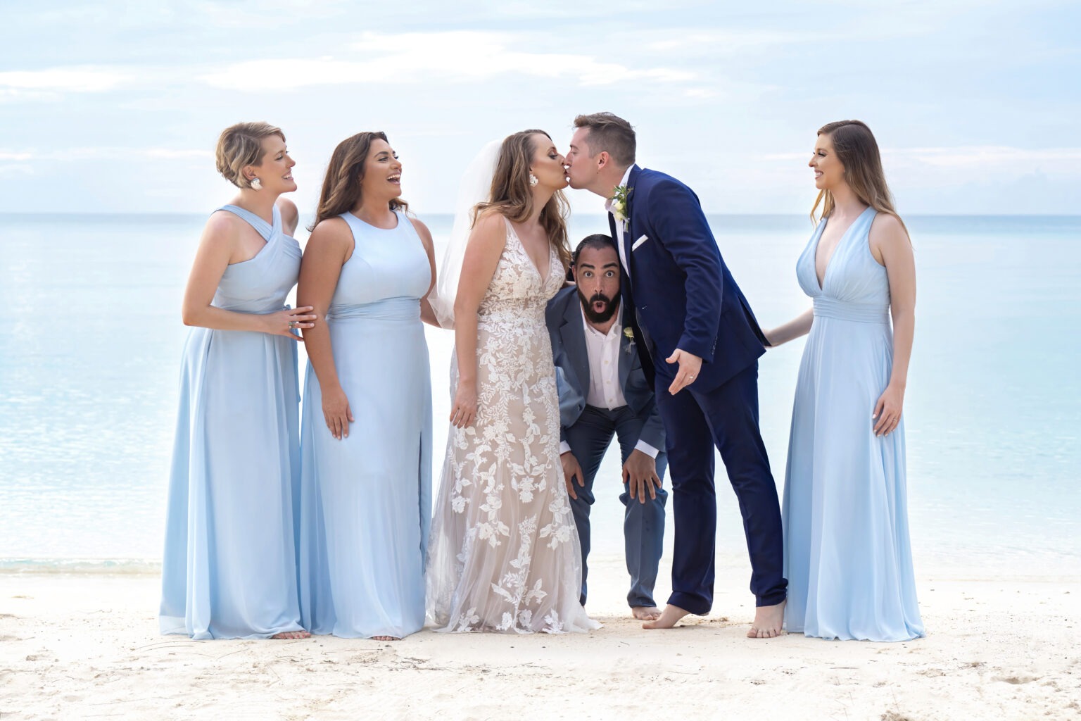 Alt Text (SEO Optimized): Playful bridesmaid photo at an Eleuthera beach wedding, featuring the bride and groom sharing a kiss surrounded by bridesmaids in soft blue dresses — and a hilarious male bridesmaid striking a fun pose. Captured by world-class destination wedding photographer David Scarola, this image radiates joy, authenticity, and island charm, showcasing his signature blend of fine-art composition and candid storytelling.