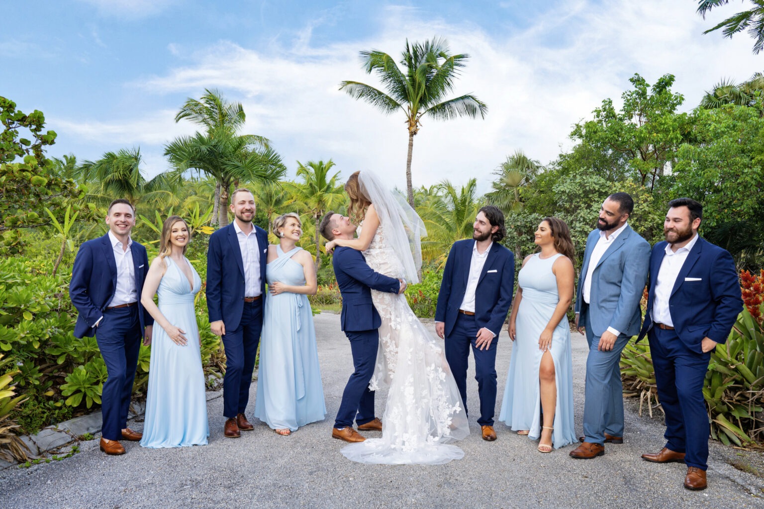 A destination wedding in Eleuthera captured through the lens of world-renowned fine art photographer David Scarola. The joyful bridal party, dressed in soft coastal blues and classic navy, stands surrounded by lush tropical palms as the newlyweds share a moment of laughter and embrace. Every detail—the golden sunlight, the textures of the island foliage, the spontaneous laughter—comes alive through Scarola’s cinematic eye and vibrant storytelling.