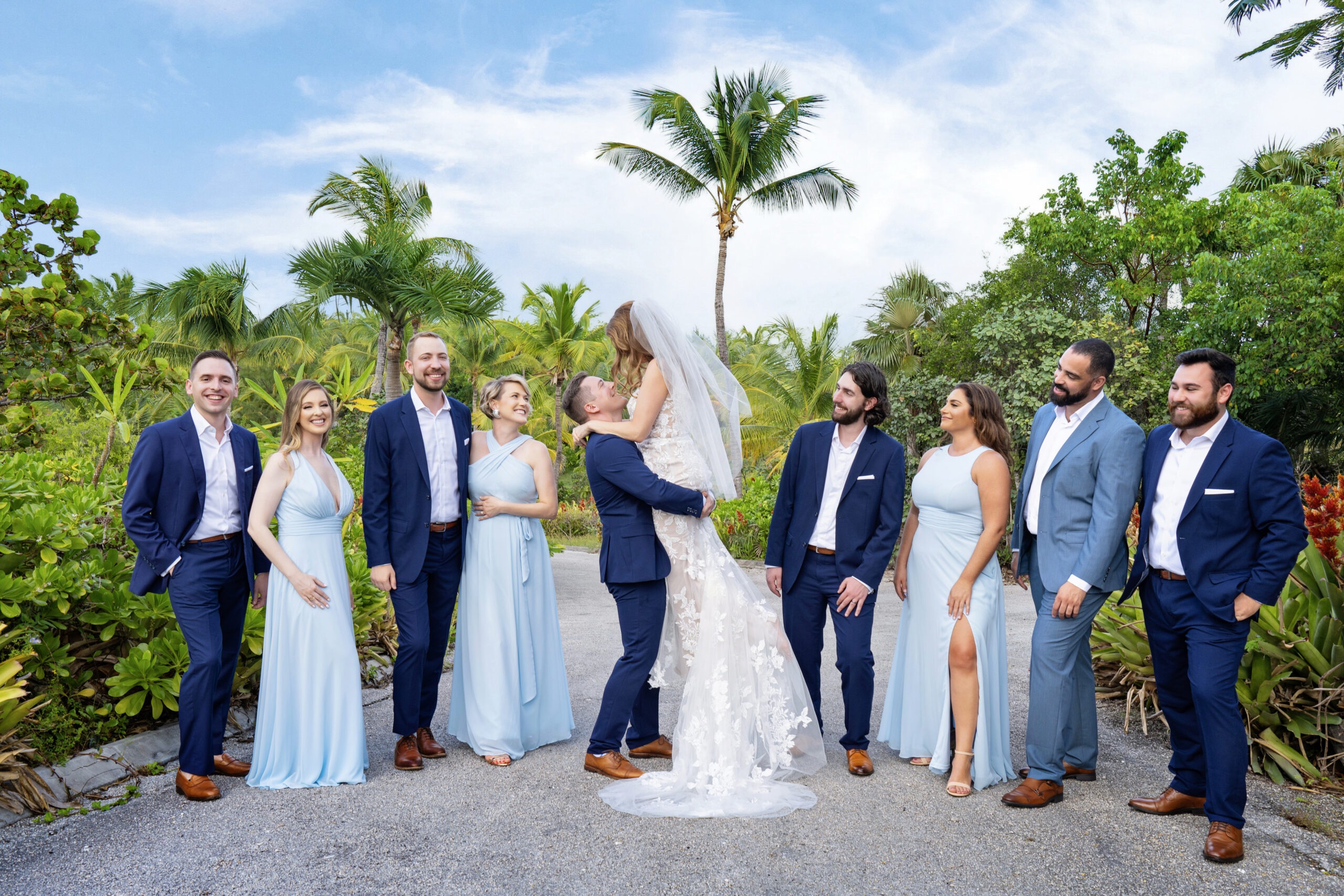 A destination wedding in Eleuthera captured through the lens of world-renowned fine art photographer David Scarola. The joyful bridal party, dressed in soft coastal blues and classic navy, stands surrounded by lush tropical palms as the newlyweds share a moment of laughter and embrace. Every detail—the golden sunlight, the textures of the island foliage, the spontaneous laughter—comes alive through Scarola’s cinematic eye and vibrant storytelling.