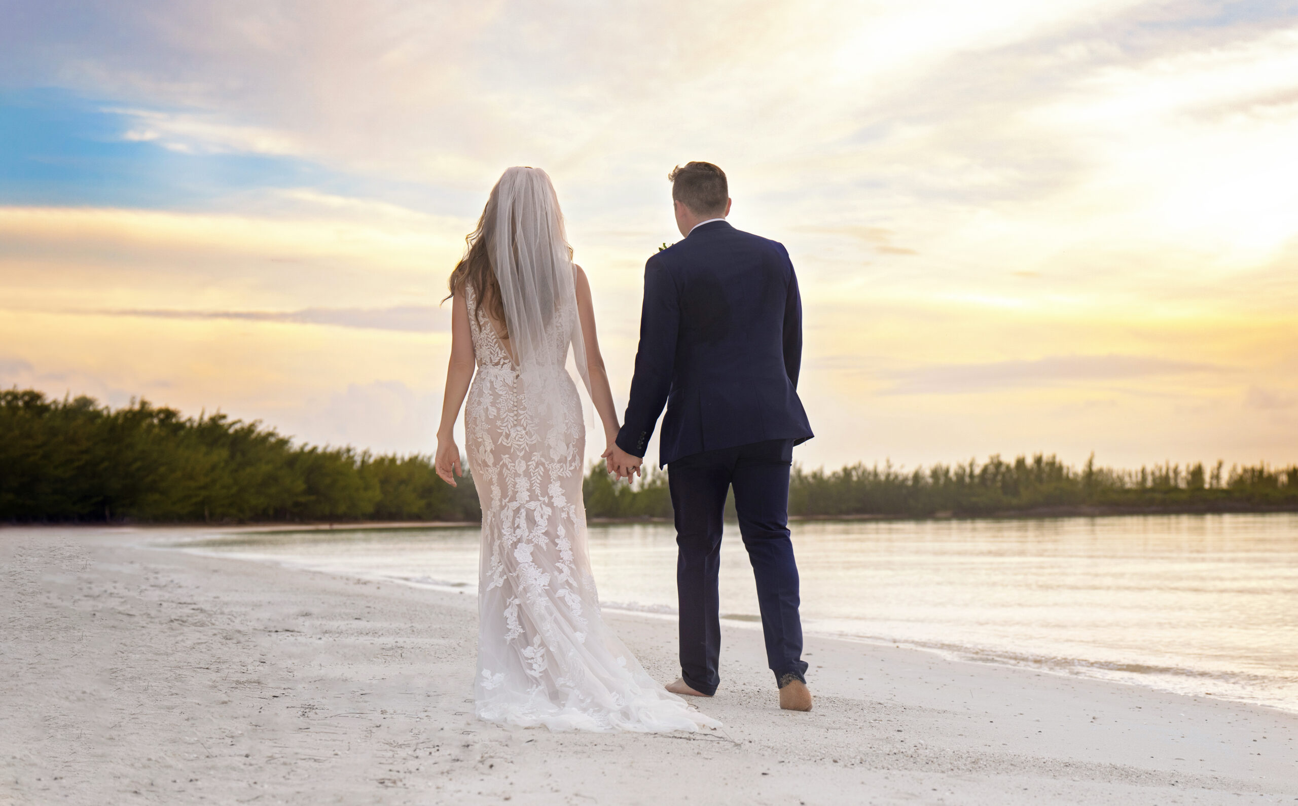 Bride and groom walking hand in hand along the shoreline at sunset in the Bahamas. The bride wears a lace gown with a flowing veil, the groom in a navy suit, both barefoot in the sand. Captured by David Scarola Photography, a fine-art wedding photographer specializing in destination beach weddings and golden-hour portraits. The sky glows with soft pastels, reflecting romance and serenity in every frame.