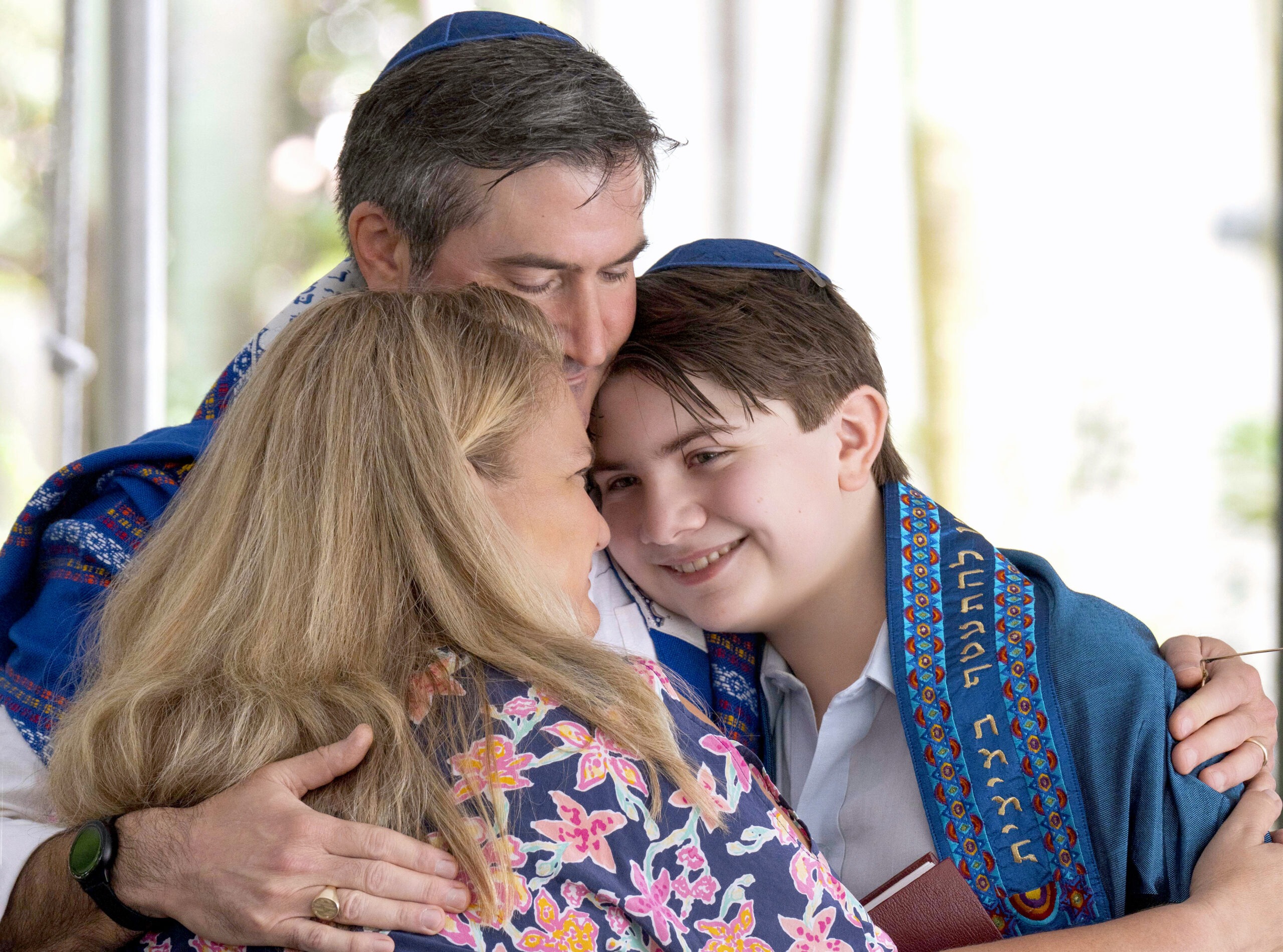 A touching family embrace during a Bar Mitzvah ceremony in Jupiter, Florida. The boy, wearing a blue tallit, smiles softly as his parents hold him close, symbolizing love, faith, and family unity.Bar Mitzvah photographer Jupiter Island, Temple Beth David events, Palm Beach event photography, luxury family portraits Florida, Jewish ceremony photographer, milestone photography Palm Beach, high-end private event photographer, David Scarola Photography, family embrace Bar Mitzvah, emotional event photography Florida, Jupiter Island photographer.