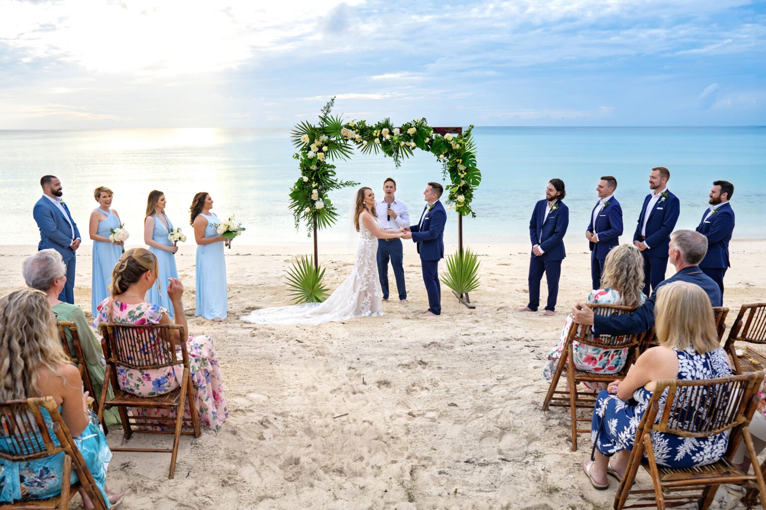A stunning Eleuthera beach wedding ceremony captured at golden hour, with the bride and groom exchanging vows beneath a lush tropical floral arch framed by the turquoise Caribbean Sea. Captured by world-class destination wedding photographer David Scarola, this image radiates warmth, authenticity, and cinematic beauty — blending fine-art composition with the natural elegance of an island sunset.