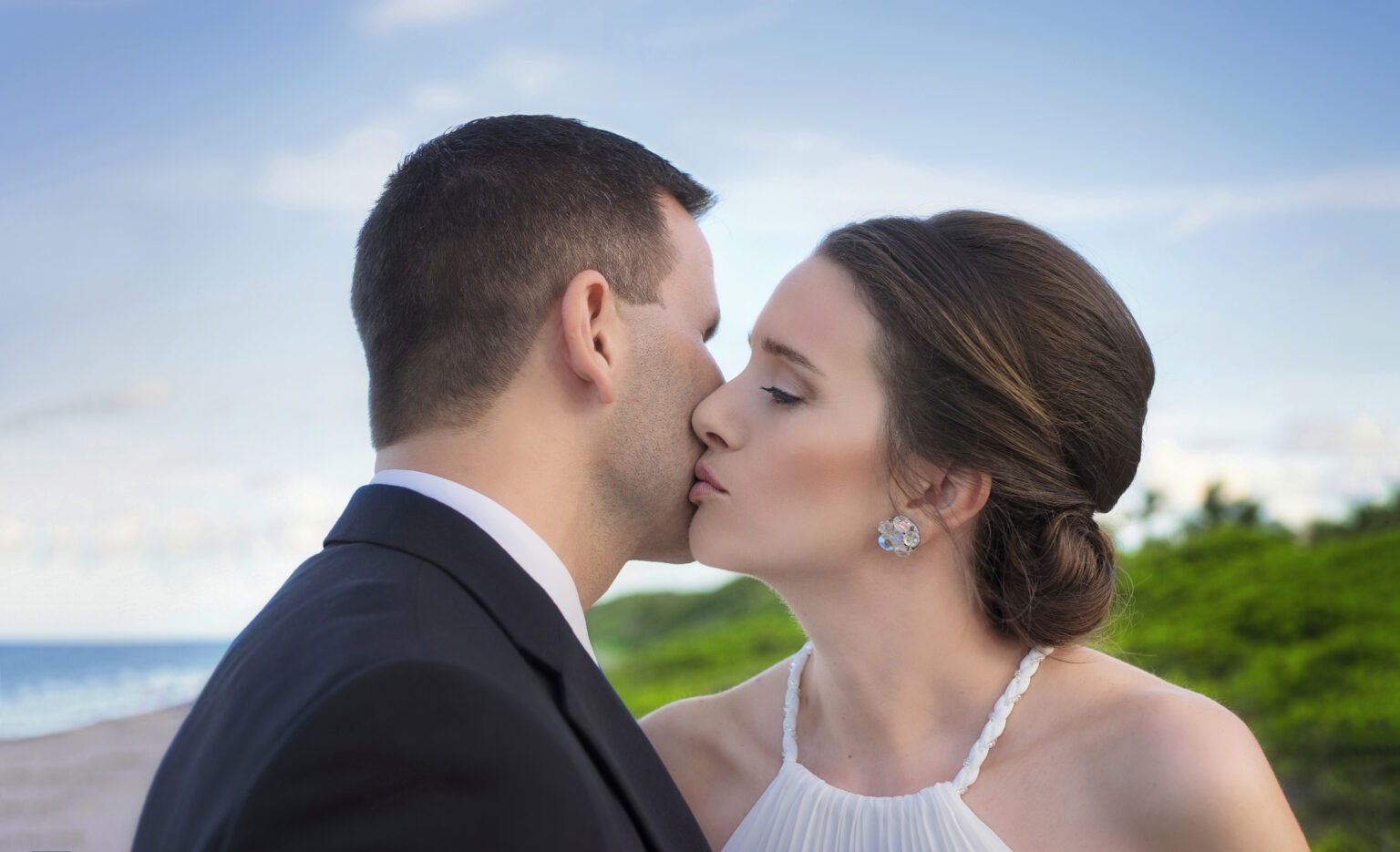 Bride and groom share an intimate kiss by the ocean at Juno Beach Pier, their love framed by the soft blue sky and coastal greenery. Captured in timeless elegance by David Scarola, Florida’s best wedding photographer and fine-art portrait artist renowned for transforming seaside weddings into cinematic, light-filled works of art.