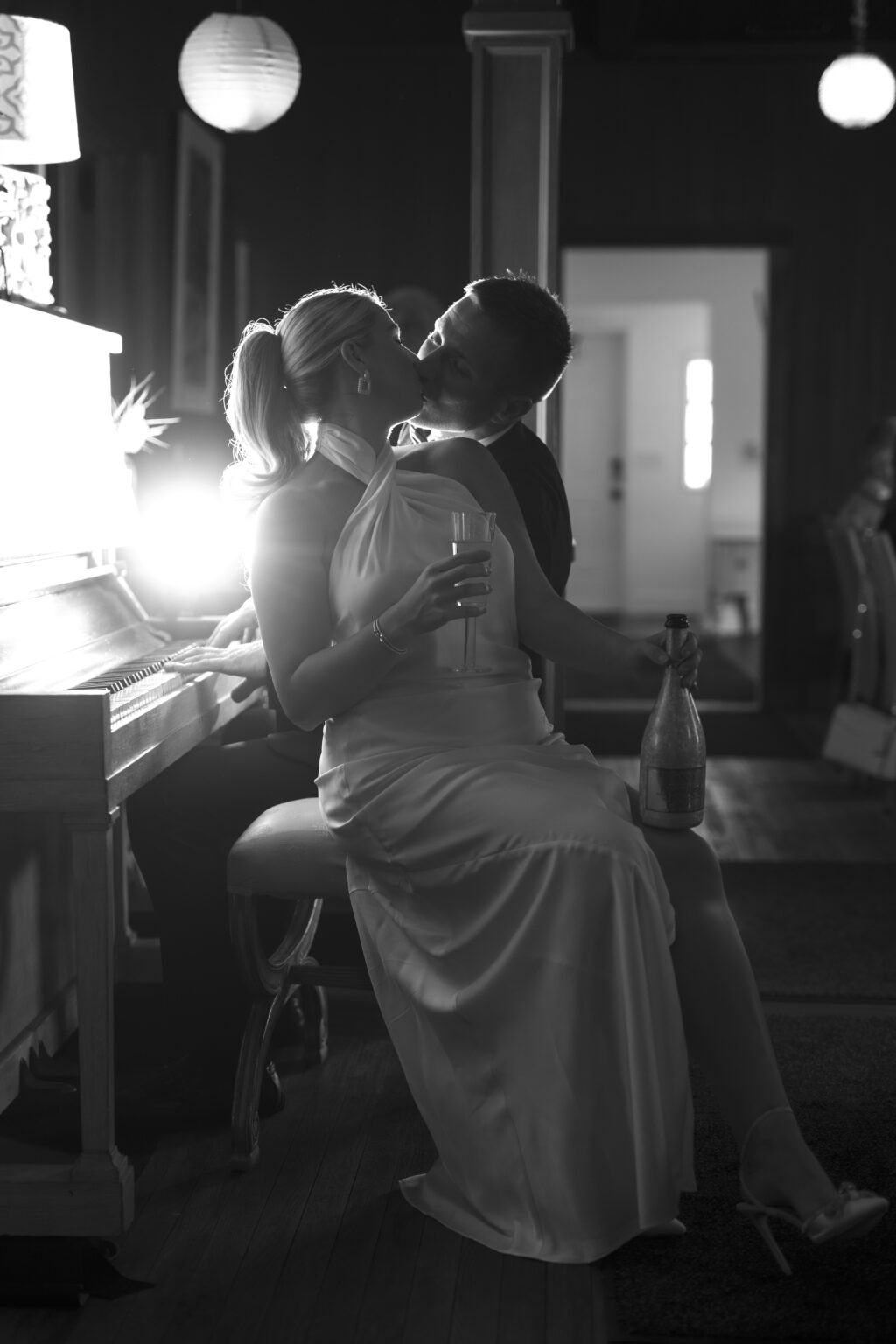 Bride and groom sharing a romantic kiss beside a softly lit piano, champagne in hand, captured in cinematic black and white — an intimate, timeless moment photographed by David Scarola, Florida’s premier wedding photographer and visionary portrait artist known for soulful, emotive, and artfully composed imagery.