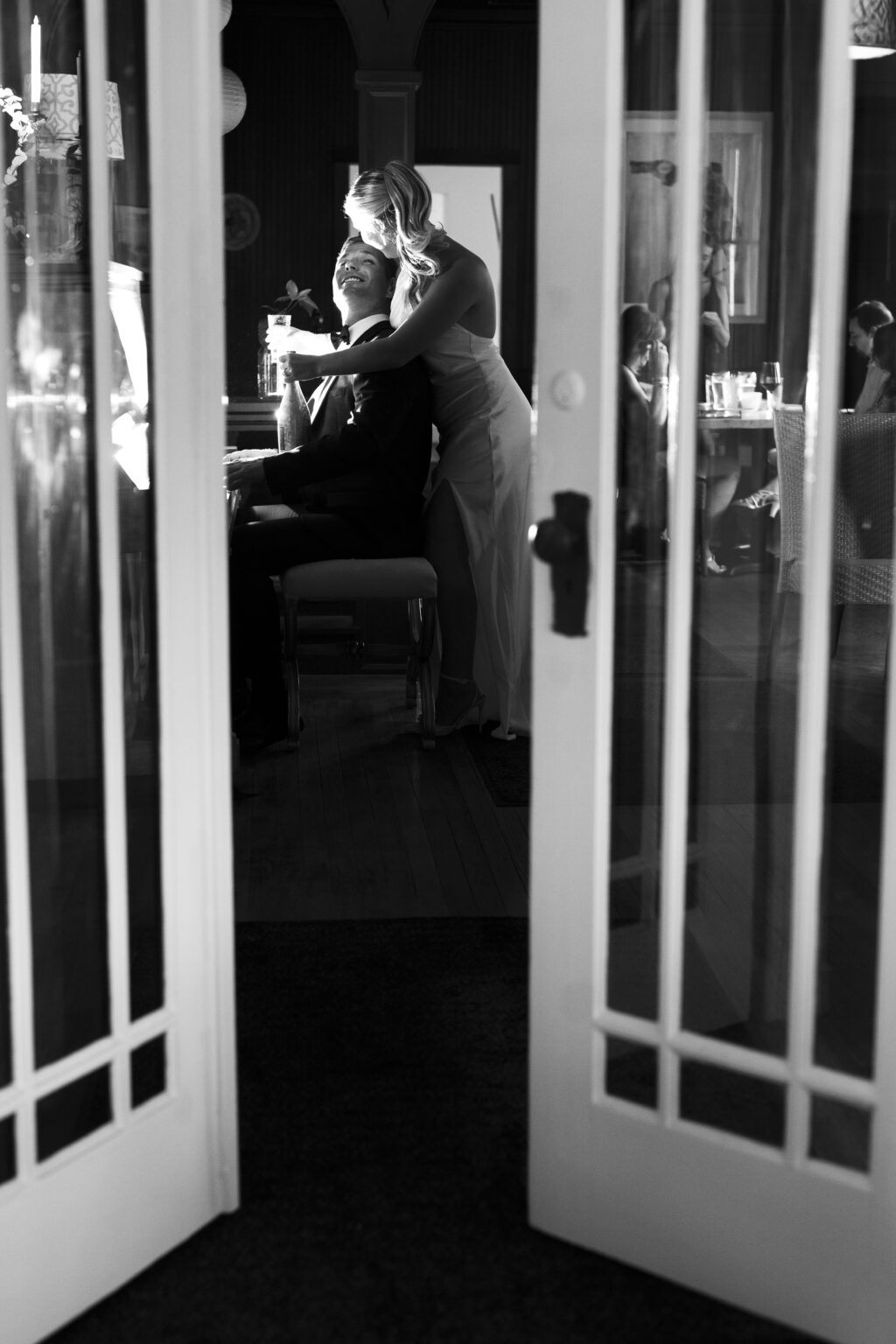 Bride and groom sharing a private, elegant moment in black and white — she embraces him from behind as he smiles at the piano with champagne in hand — captured in timeless fine-art style by David Scarola, Palm Beach’s best wedding photographer and master of classic black-and-white portraiture.