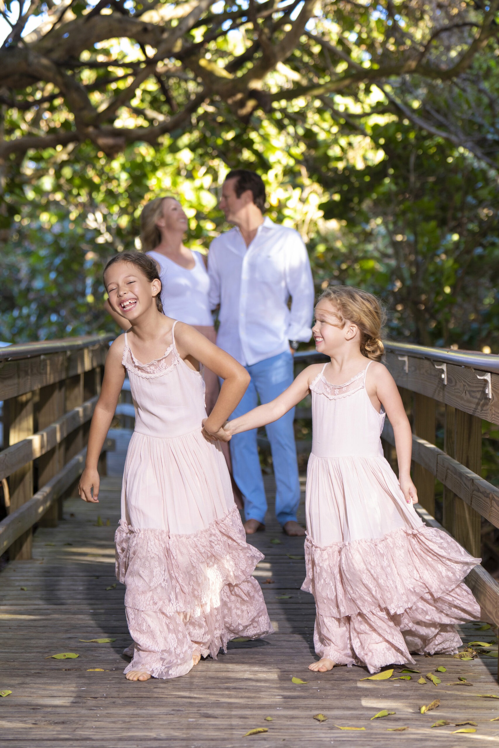 Sisters laughing and running hand in hand on a boardwalk surrounded by lush greenery, with parents smiling in the background during a family photo shoot on Jupiter Island, captured by master portrait photographer David Scarola.Jupiter Island family photography, Palm Beach County photographer, fine art family portraits, lifestyle family session, natural light photography, children laughing, candid family portraits Florida, David Scarola Photography, master portrait photographer, Palm Beach family beach photography