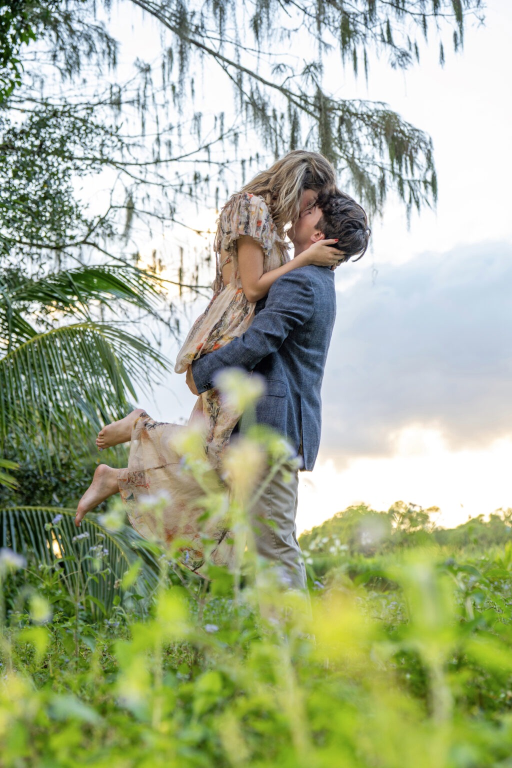 Romantic engagement photo of Lucy and Brody sharing a kiss outdoors during golden hour, captured in lush greenery by David Scarola Photography. A natural, heartfelt portrait symbolizing love and connection — ideal for engagement and wedding photography in South Florida.engagement photographer Jupiter FL, Palm Beach wedding photography, romantic couples photography Florida, fine art engagement portraits, outdoor engagement photo shoot, South Florida lifestyle photographer, top engagement photographer Palm Beach County, natural light wedding portraits, David Scarola Photography, Jupiter Island engagement photographer.